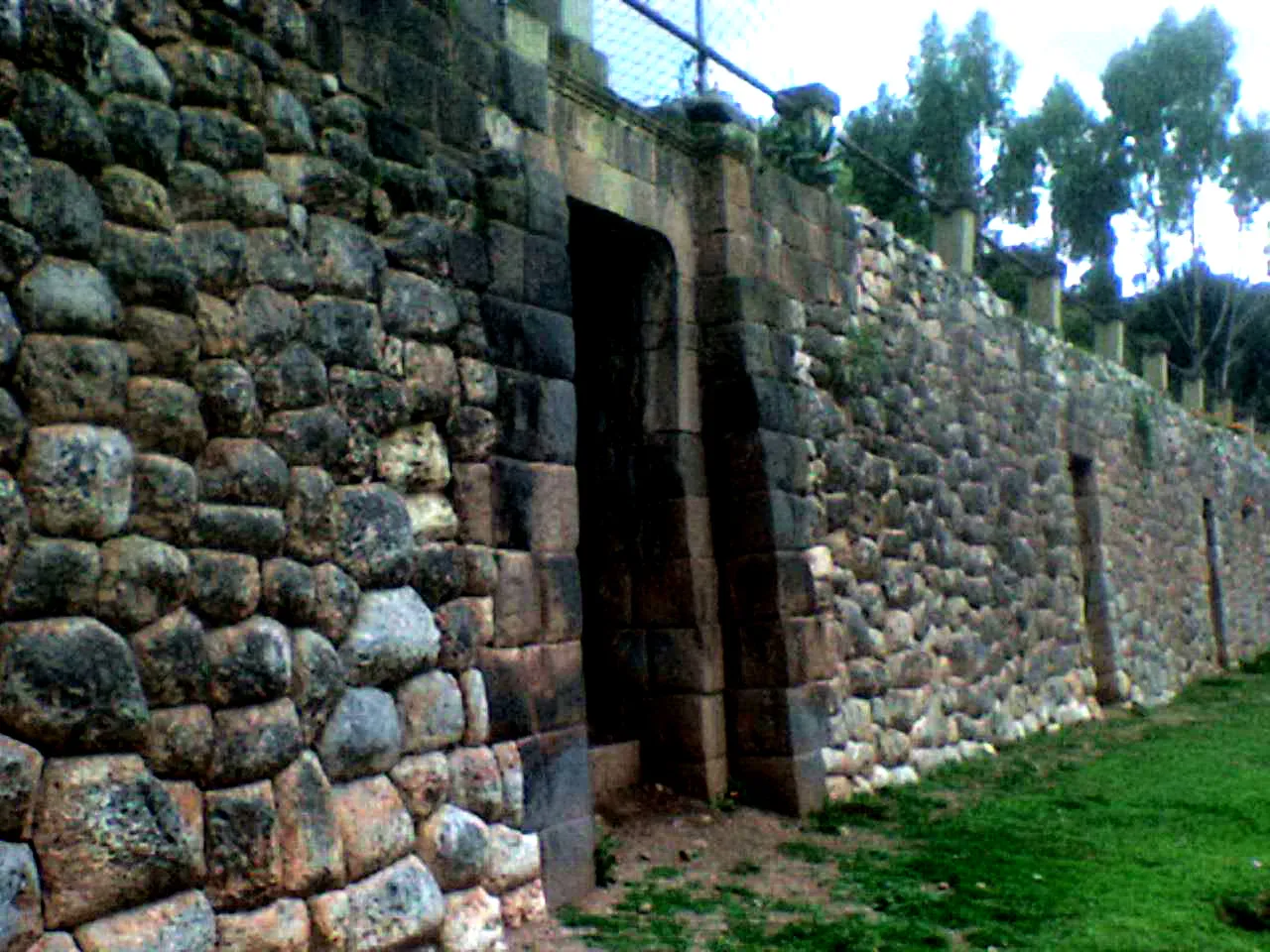 The Inca wall at Colcampata in Cusco, built from irregular interlocking stones with a tall trapezoidal doorway cut through it and grass along its foot