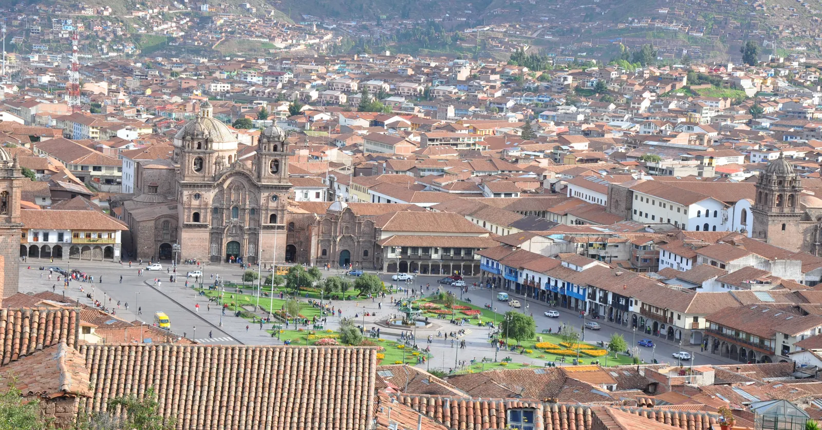Cusco's Plaza de Armas seen from the Colcampata terrace above it, with the twin towers of the Jesuit church, the arcaded shopfronts along two sides and terracotta roofs spreading beyond