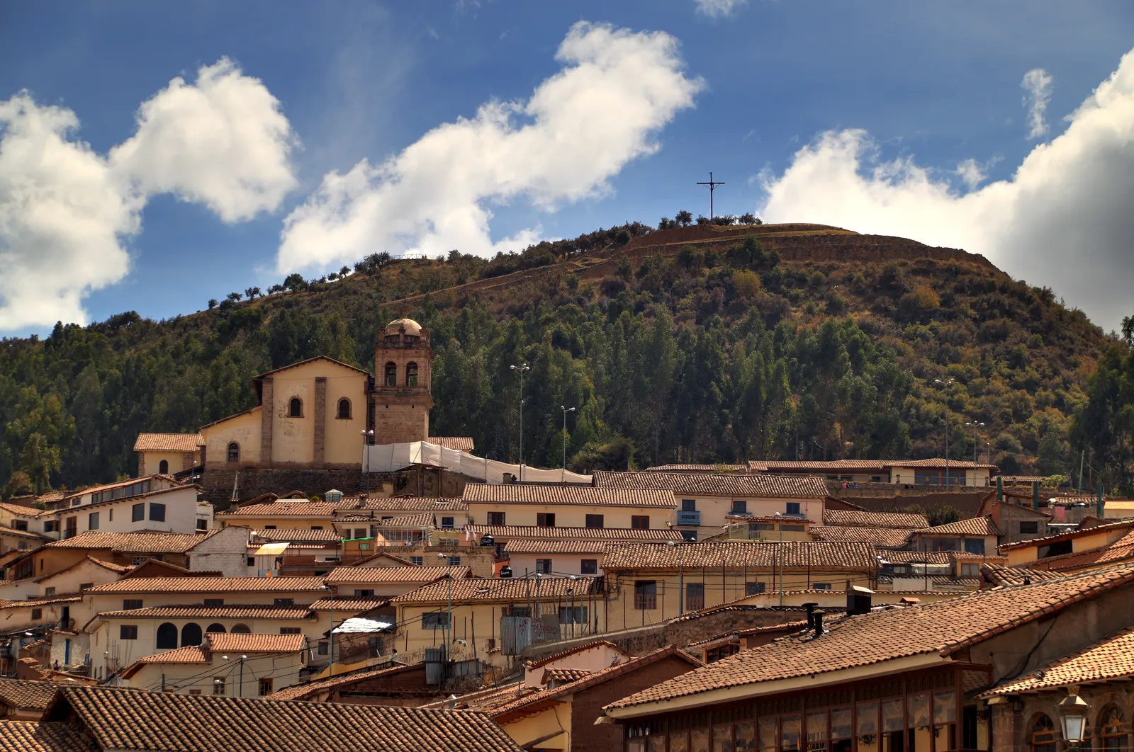 The church of San Cristóbal in Cusco with its square bell tower, standing on the hillside above the terracotta roofs of the old town, a hilltop cross on the slope behind it