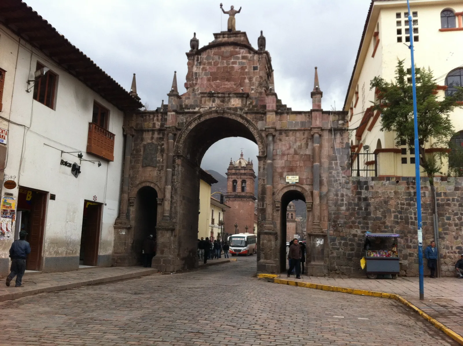 The Santa Clara arch in Cusco, a three-arched stone gateway with a figure on top, spanning a cobbled street with a church tower visible through the central opening