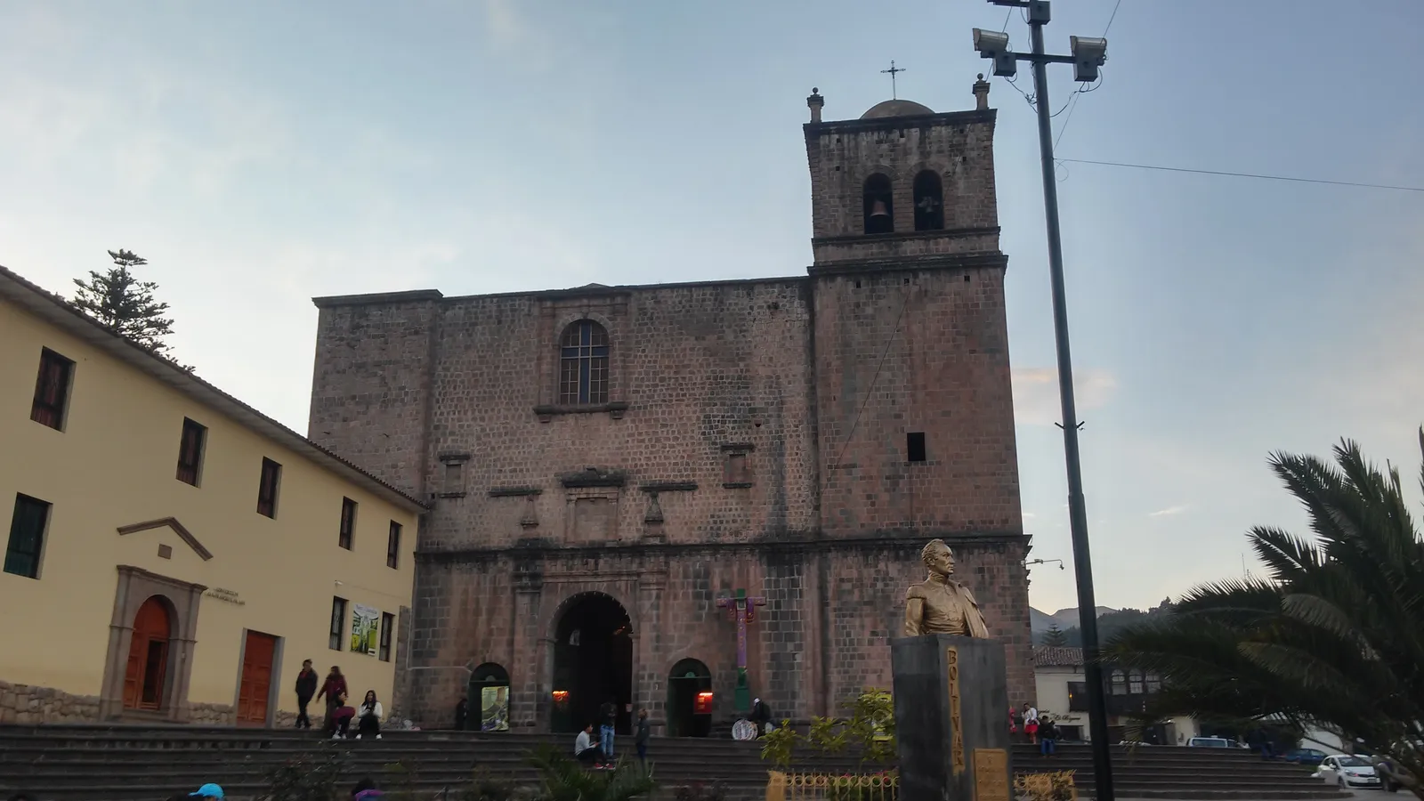 The stone facade and single bell tower of the church of San Francisco de Asís in Cusco, seen from the steps of Plaza San Francisco with people crossing in front of it