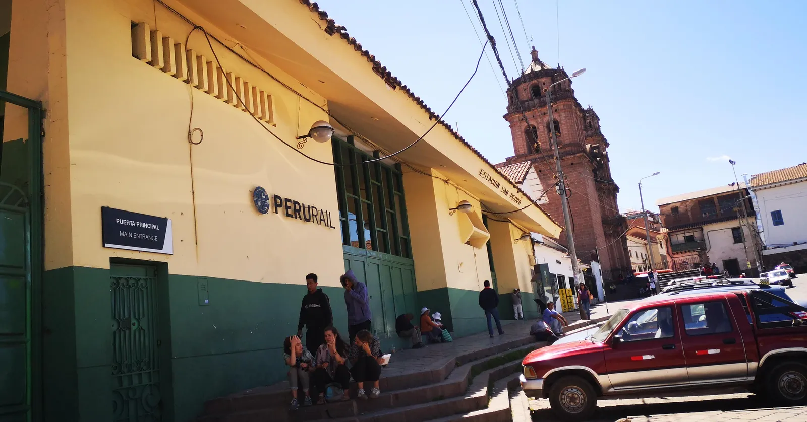 The yellow and green street frontage of San Pedro station in Cusco, with its arched green doors open onto the pavement and the bell tower of the San Pedro church rising behind it