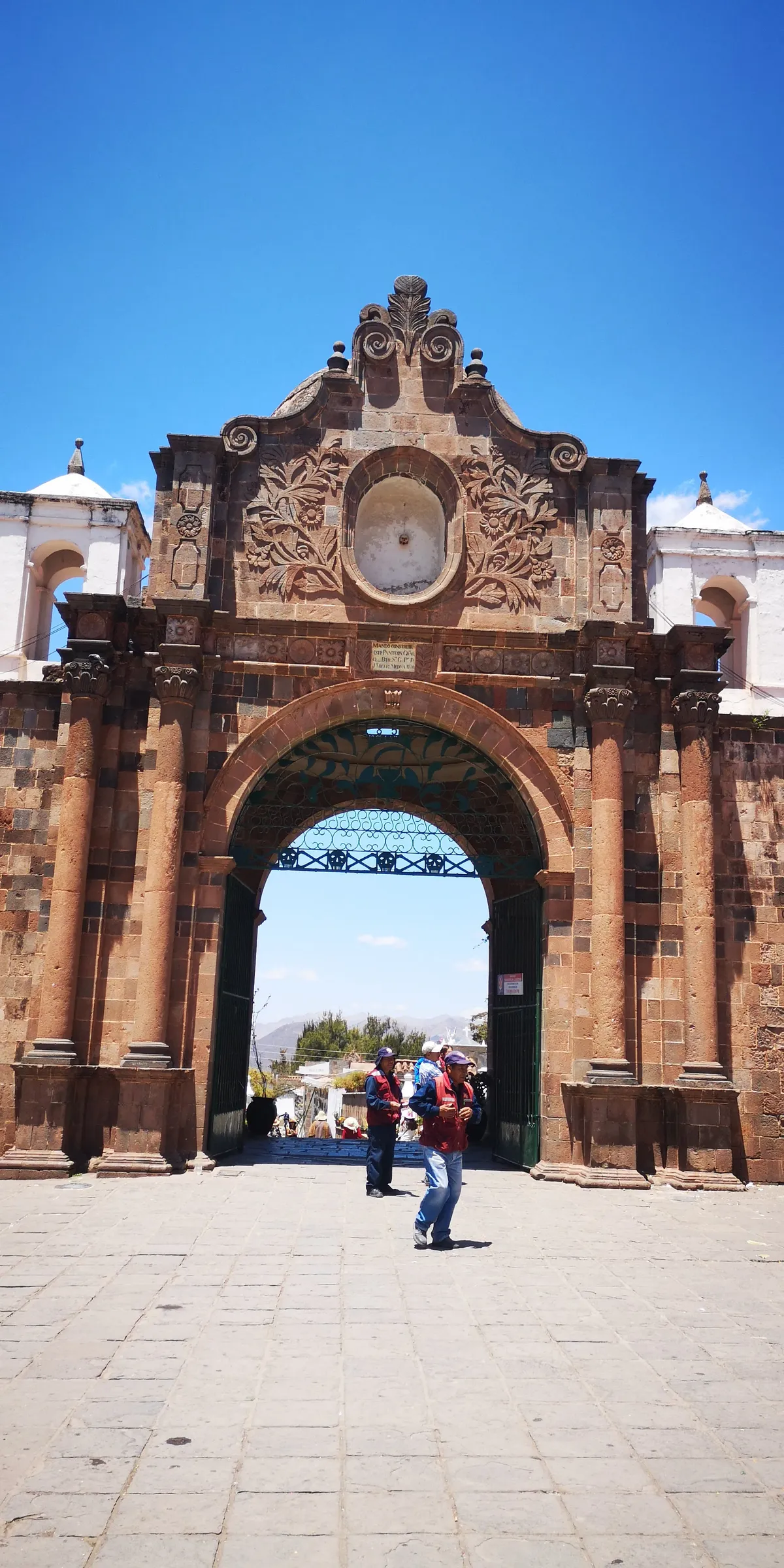 The carved sandstone gateway of the Almudena cemetery in Cusco, a tall arch with foliage carving and an oval niche above it, open onto the graves beyond
