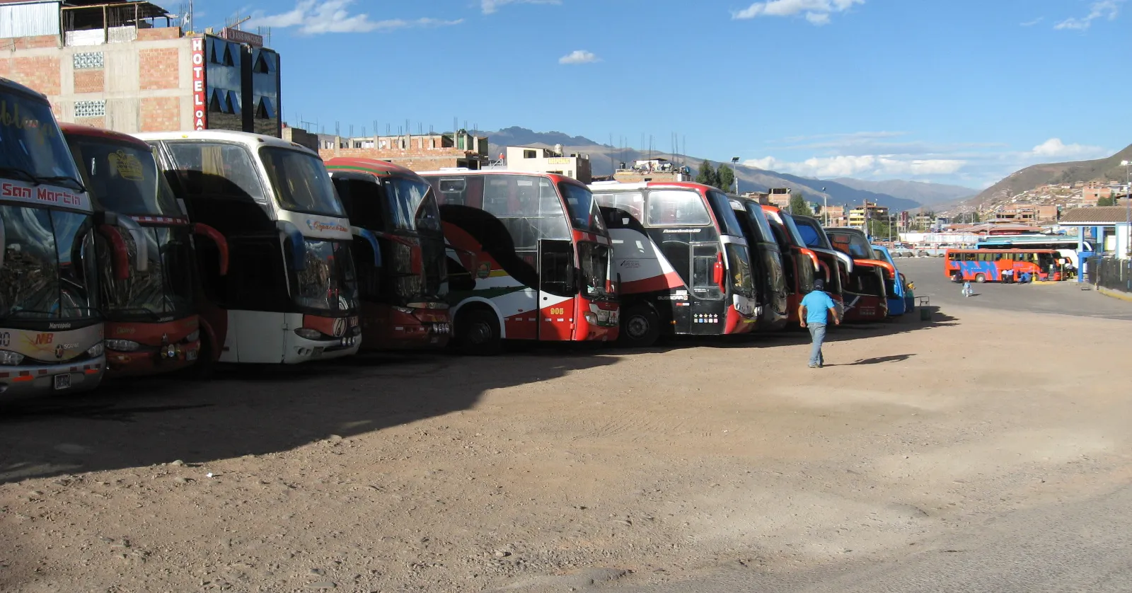 A line of double-decker long-distance coaches parked nose-out on the gravel apron at Cusco's Terminal Terrestre, with the city's hillsides in the distance