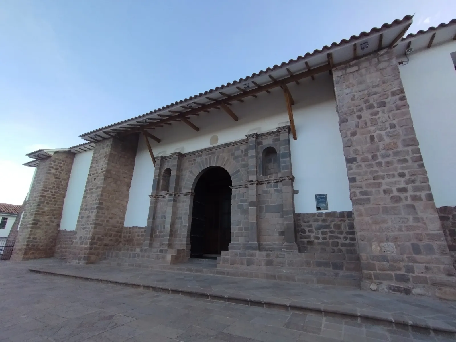 The front of the Templo de Santiago Apóstol in Cusco, a whitewashed church with stone buttresses, a carved stone doorway under a tiled porch roof, and a paved square in front of it