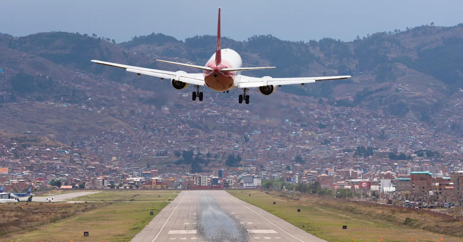 An airliner on short final over the runway at Cusco's airport, with the city's rooftops and the brown Andean hillsides filling the background