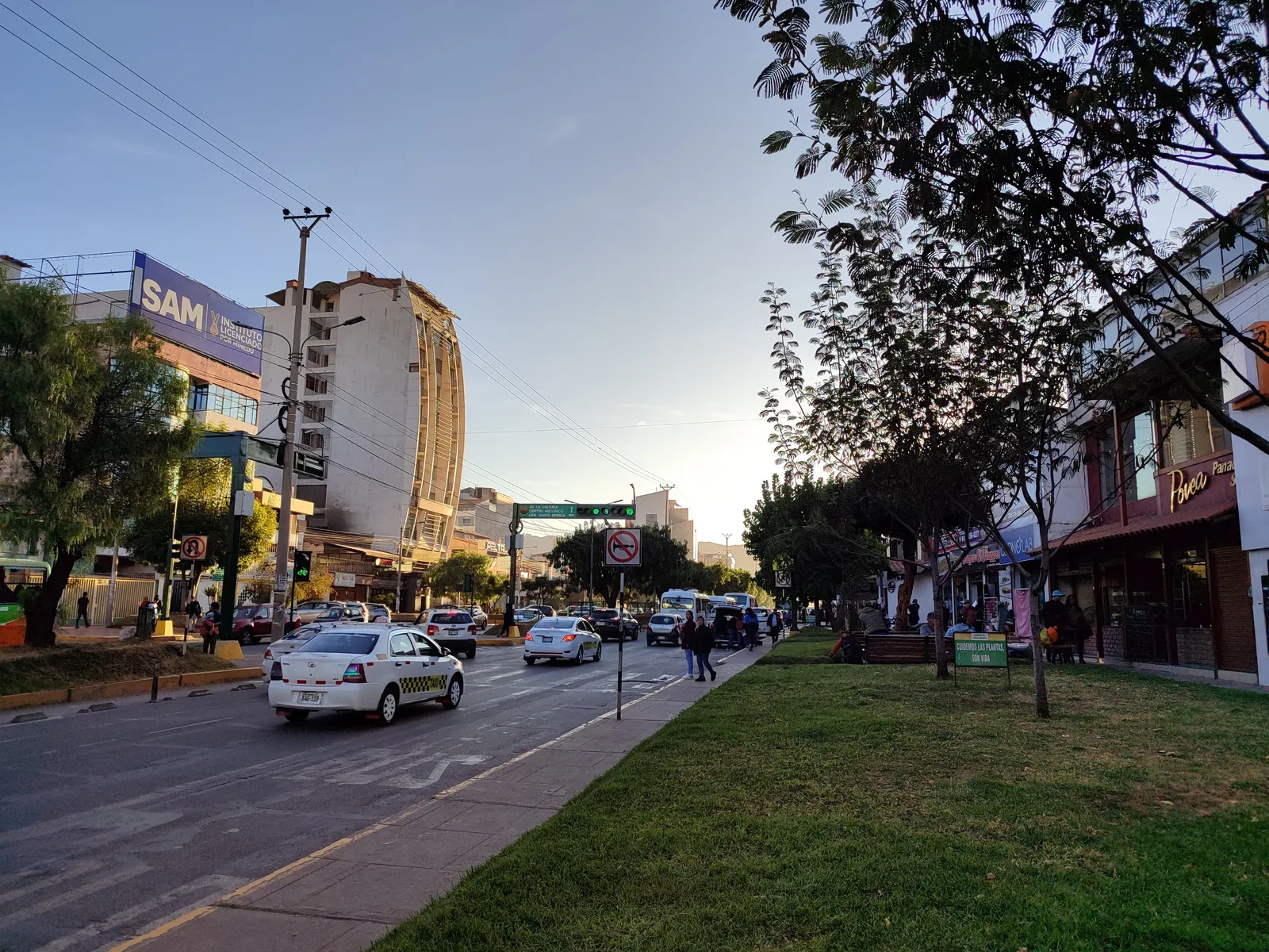 Avenida de la Cultura in Cusco in late afternoon, taxis and buses on the carriageway with a planted central reservation and shopfronts along the pavement