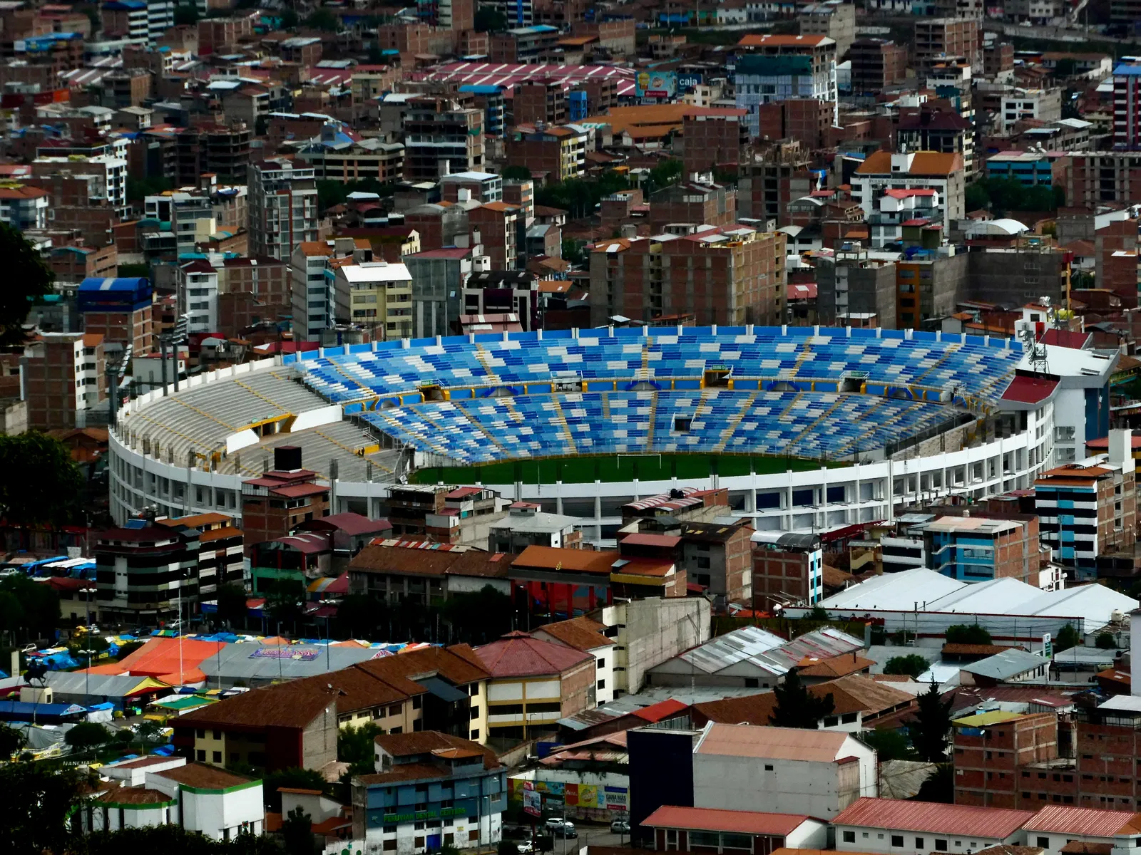 The Estadio Inca Garcilaso de la Vega in Cusco seen from the hillside above, its blue seating bowl surrounded on every side by the tin and tile roofs of the city