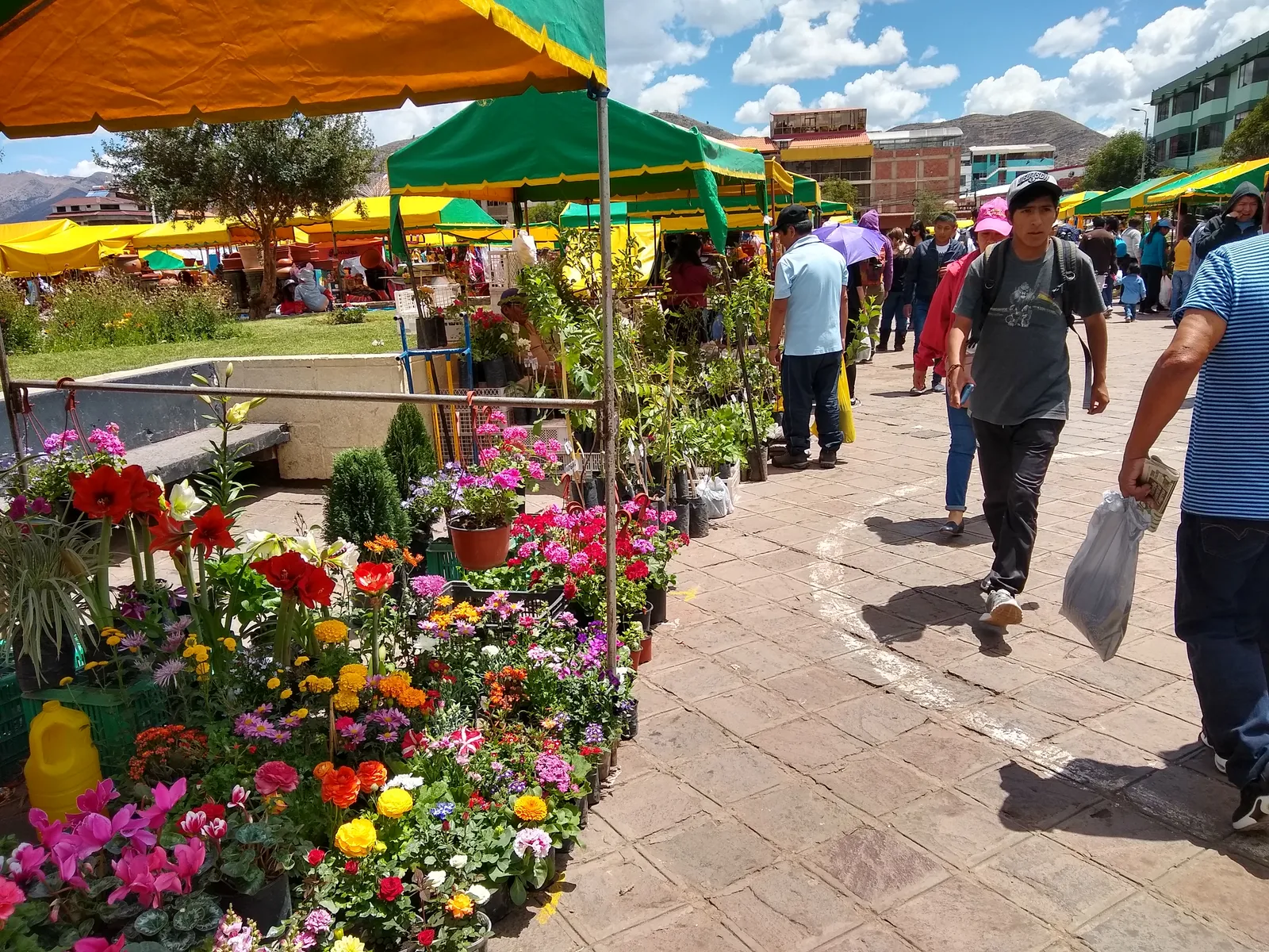 Flower stalls under green and yellow canopies on the paved square at Plaza Túpac Amaru in Cusco, with shoppers walking past carrying bags