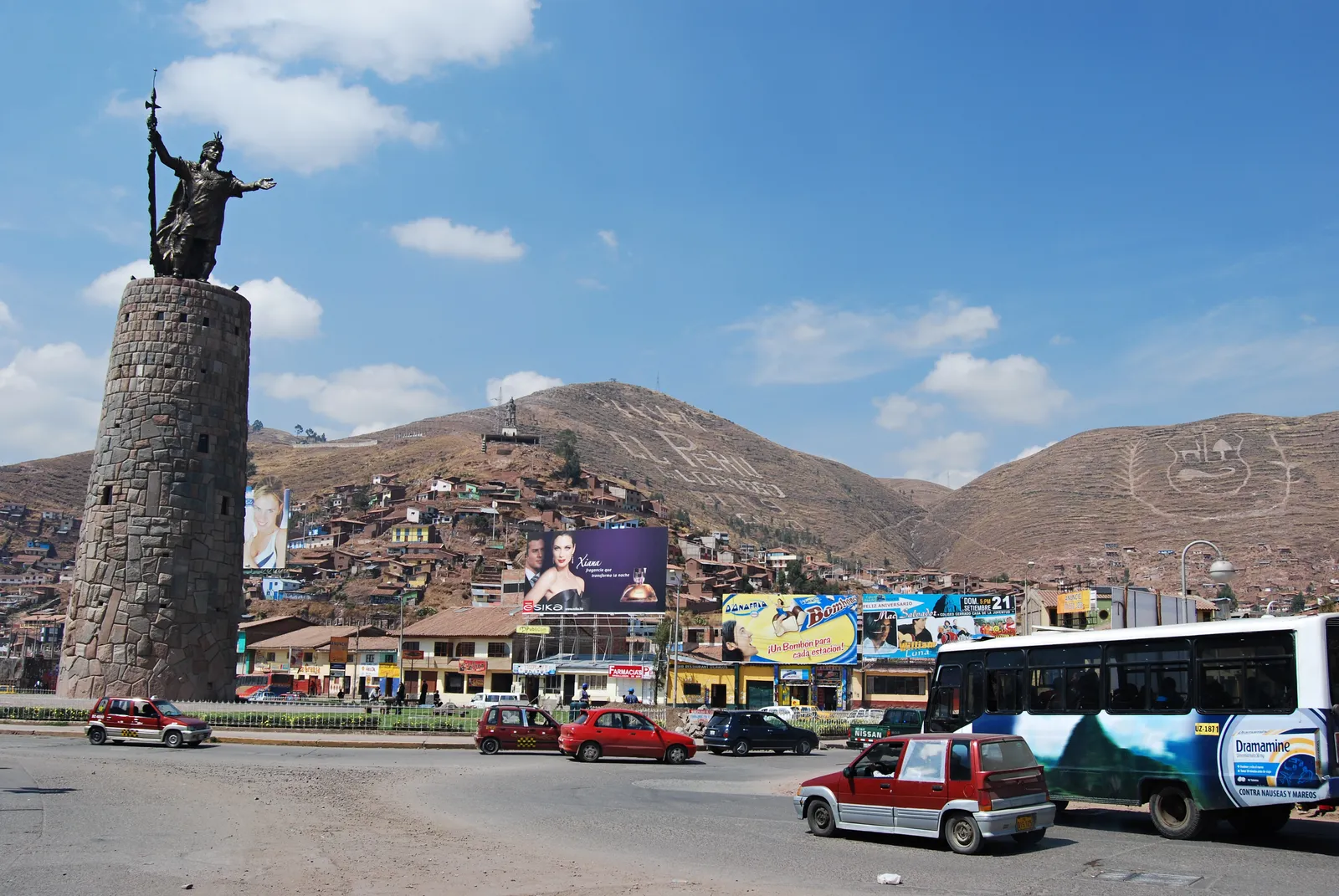 The Pachacútec monument in Cusco, a bronze figure with a raised staff standing on a round stone tower in the middle of a traffic roundabout, with the hillside houses of the city behind