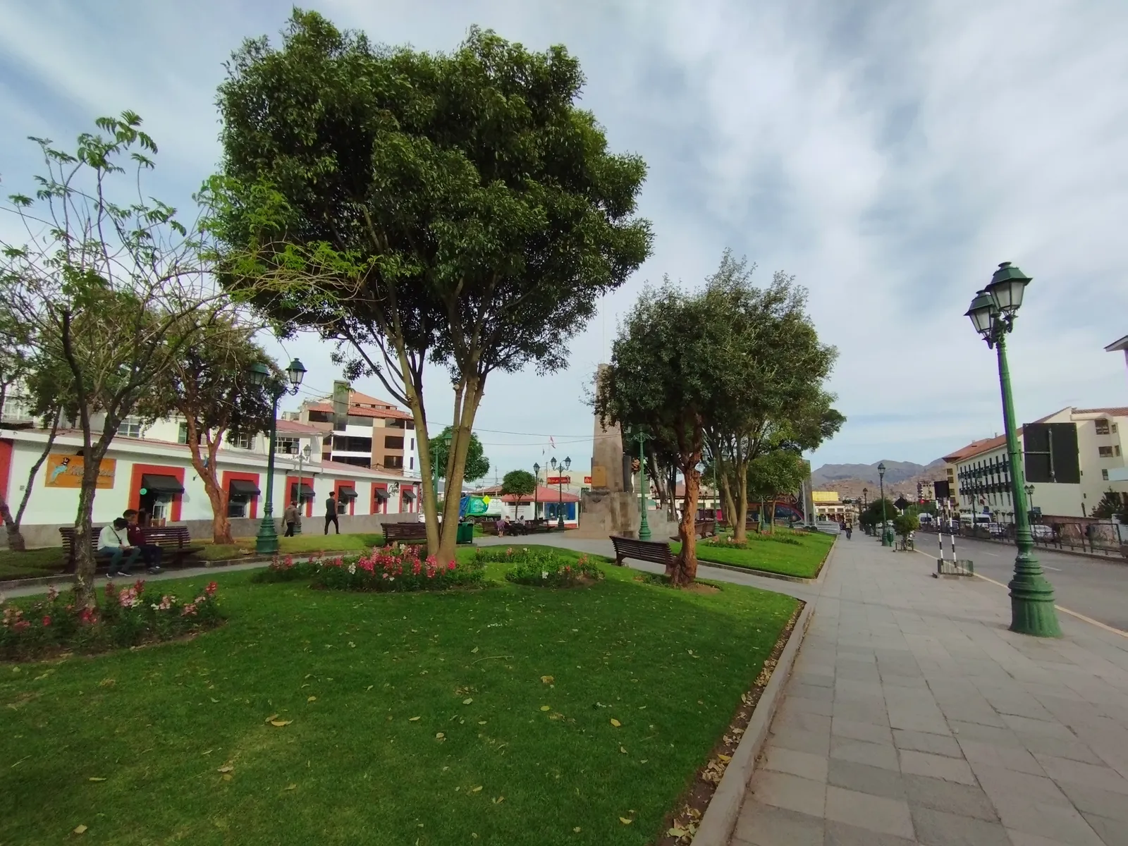 Parque Orellana at Pumaqchupan in Cusco, a lawn and young trees beside a wide paved promenade with benches, low shopfronts along one side and hills in the distance