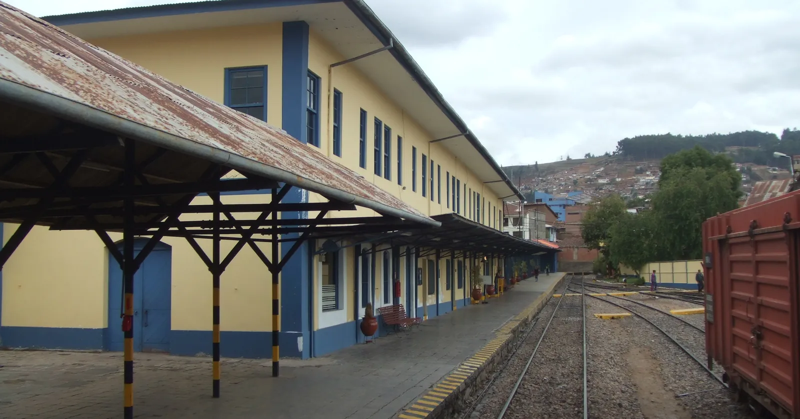 The single platform at Wánchaq station in Cusco, with the yellow and blue station building under a long canopy on rusted iron posts and the hillside city rising beyond the tracks