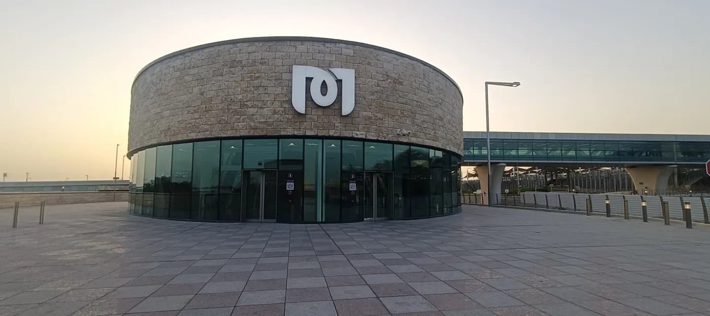 The stone drum of the Hamad International Airport metro station entrance, glazed at ground level with the Doha Metro symbol on the wall above the doors