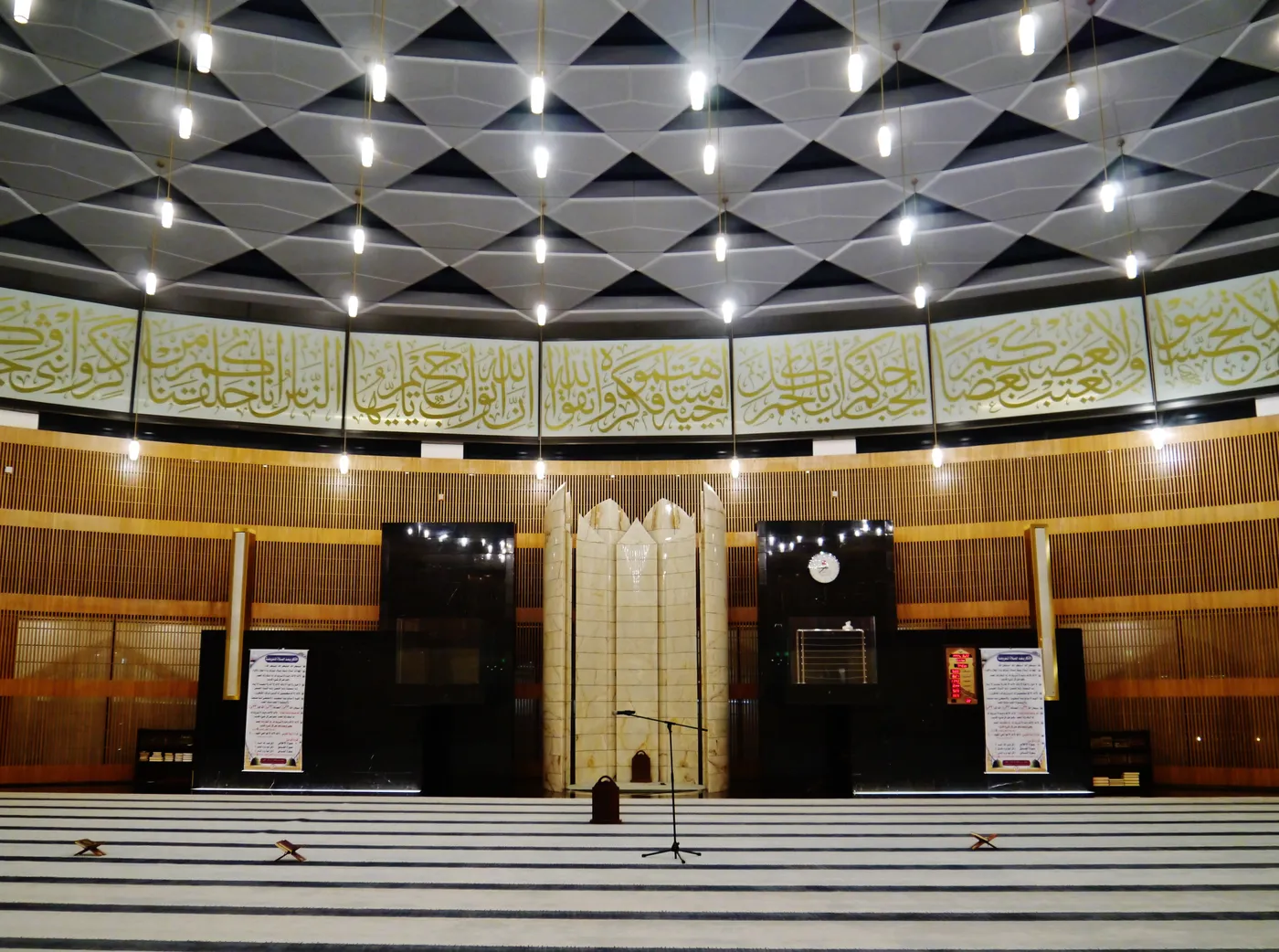 The prayer hall of the mosque at Hamad International Airport, a wide carpeted floor facing a pale stone mihrab under a faceted ceiling with a band of Arabic calligraphy running around the walls