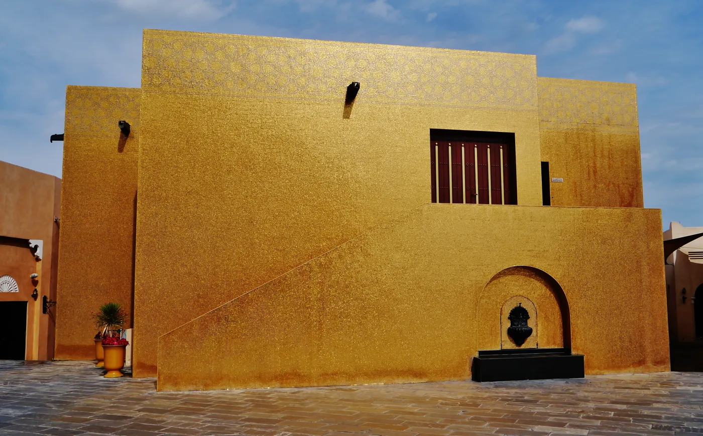 The Golden Mosque at Katara Cultural Village in Doha, a plain cubic building clad from ground to roofline in small gold mosaic tiles, with a dark fountain niche set into one wall