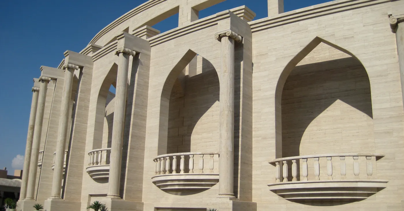 The curving travertine outer wall of the amphitheatre at Katara Cultural Village in Doha, deep pointed-arch niches with stone balconies set into it and benches along the paving below