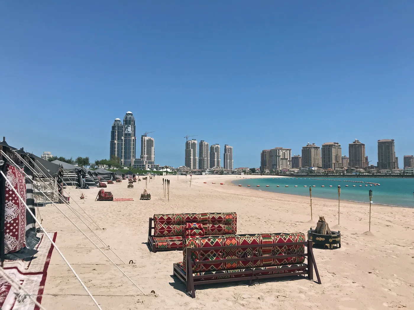 Katara Beach in Doha, patterned majlis benches set on pale sand above a turquoise shallows, with the towers of the Pearl-Qatar on the horizon