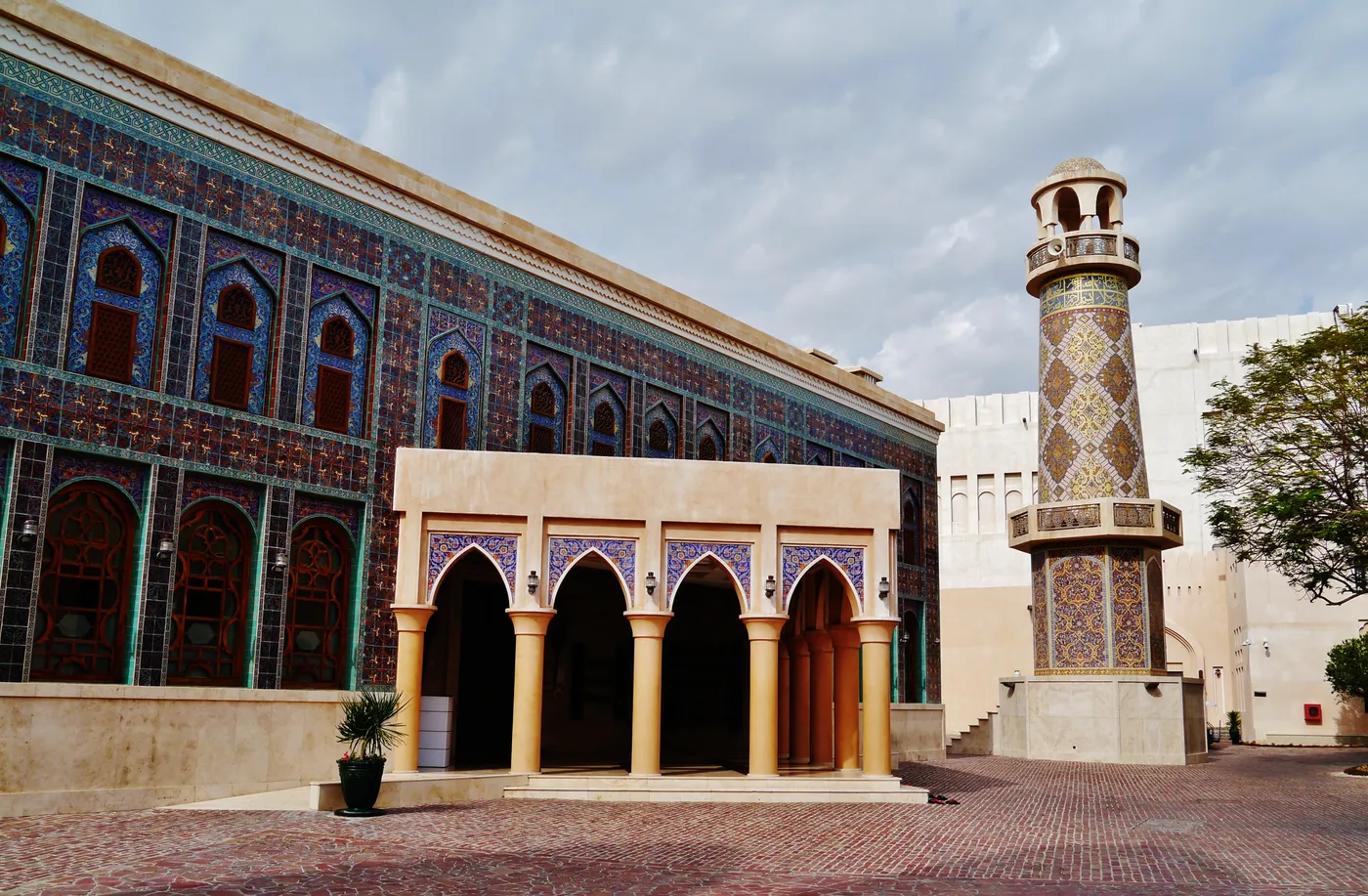 The Katara Mosque in Doha, its walls and arcade faced in turquoise and gold tilework, with a cylindrical tiled minaret standing beside it over a brick-paved courtyard