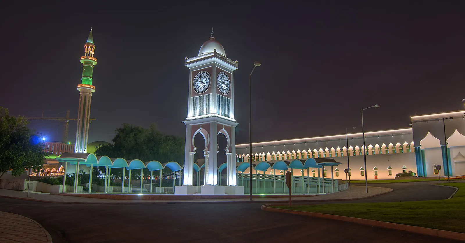 The white clock tower in front of the Amiri Diwan in Doha, floodlit at night, with a green-capped minaret behind it and a long arcaded wall to the right