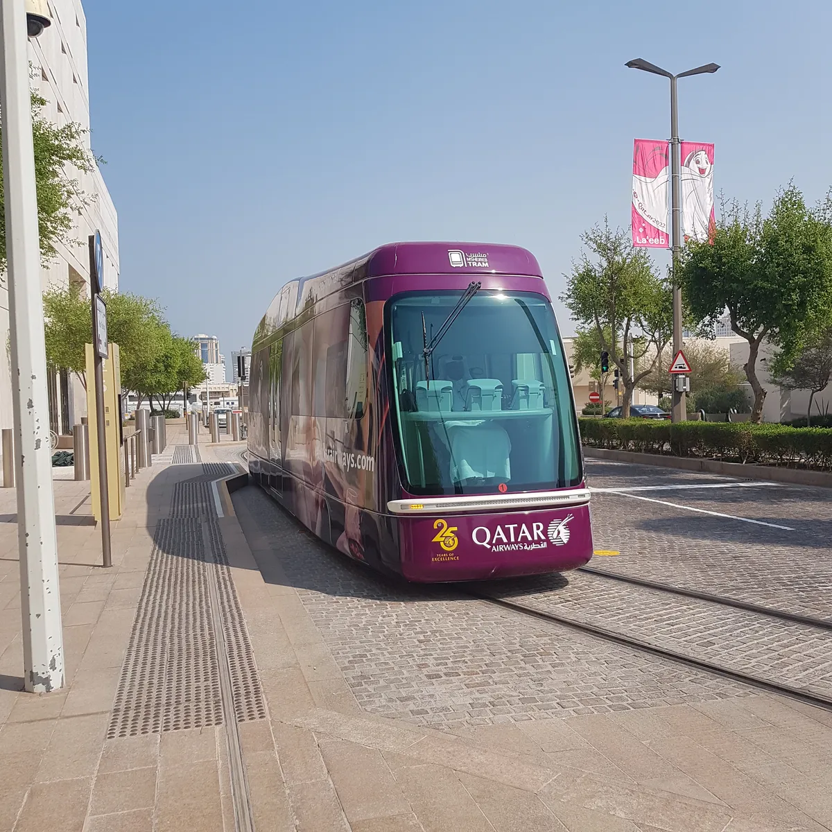 A purple Msheireb tram stopped on street-level tracks in Doha, with paving and young trees along the kerb and a flag banner on a lamppost behind