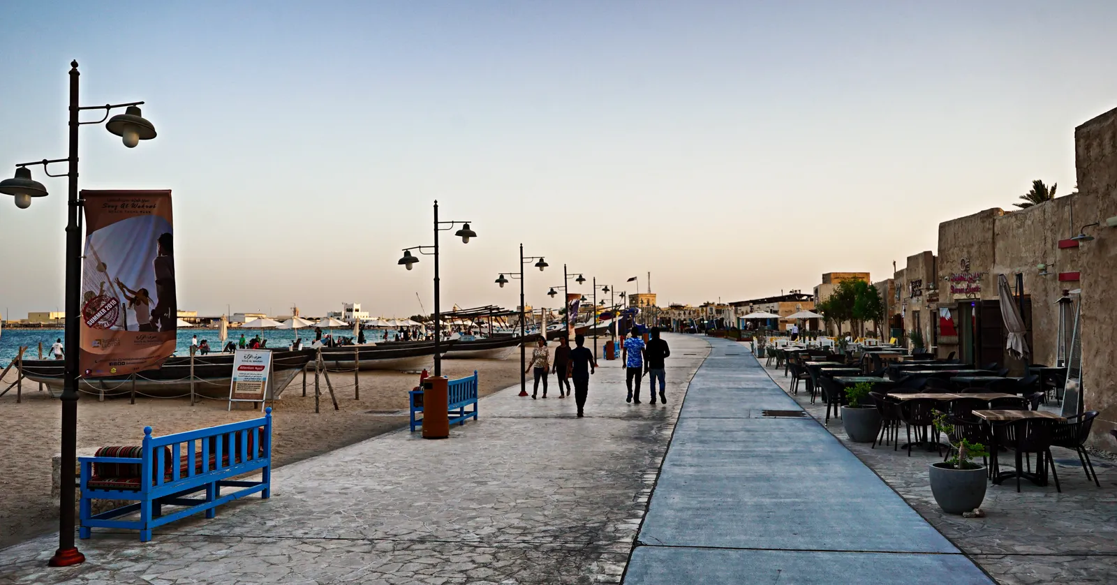 The seafront boardwalk at Souq Al Wakrah at dusk, restaurant terraces along low sandstone buildings on the right and boats drawn up on the sand to the left