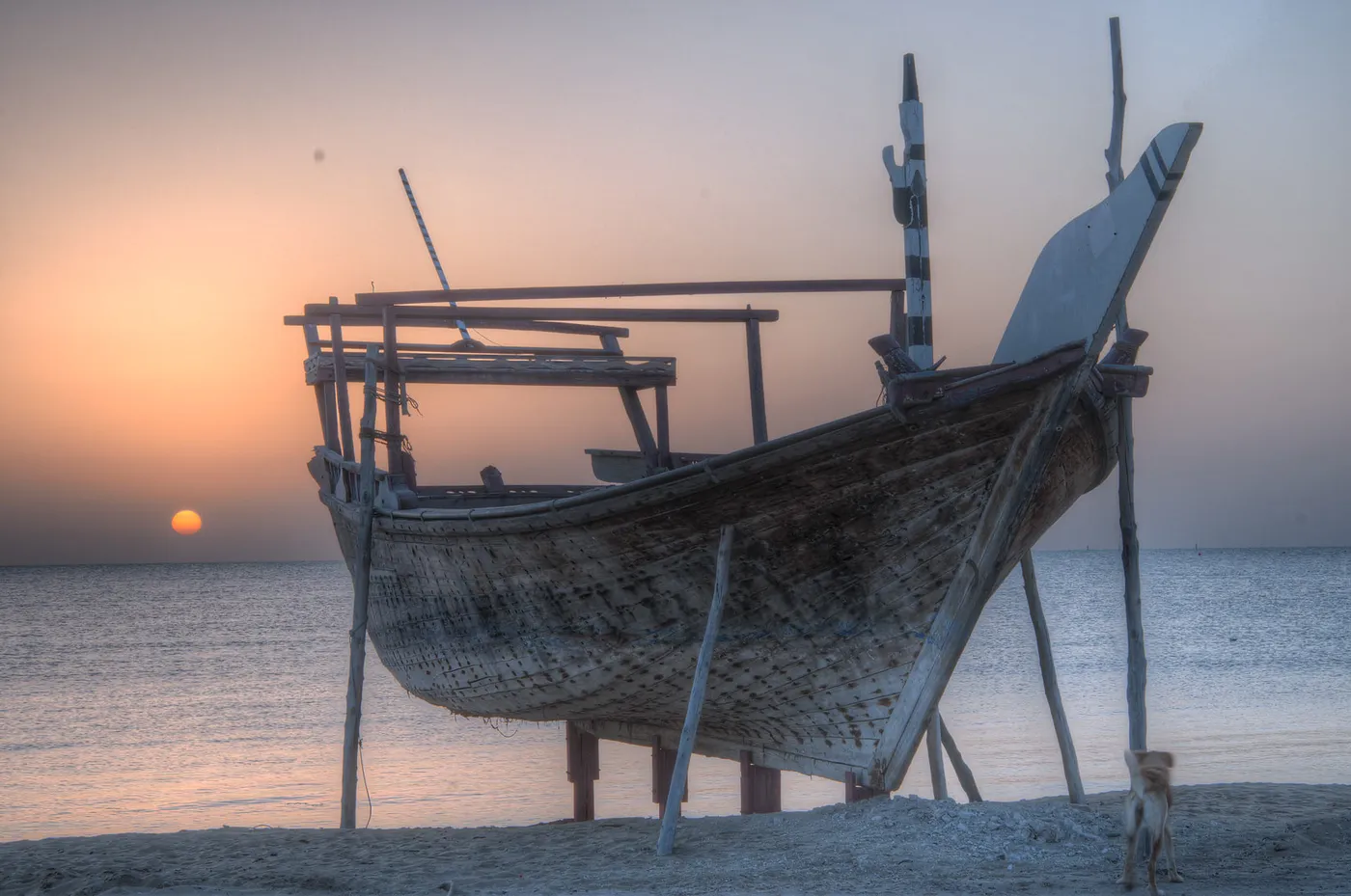 A wooden fishing dhow propped upright on timber legs on the sand at Al Wakrah, its hull and rigging dark against a hazy sunrise over the water