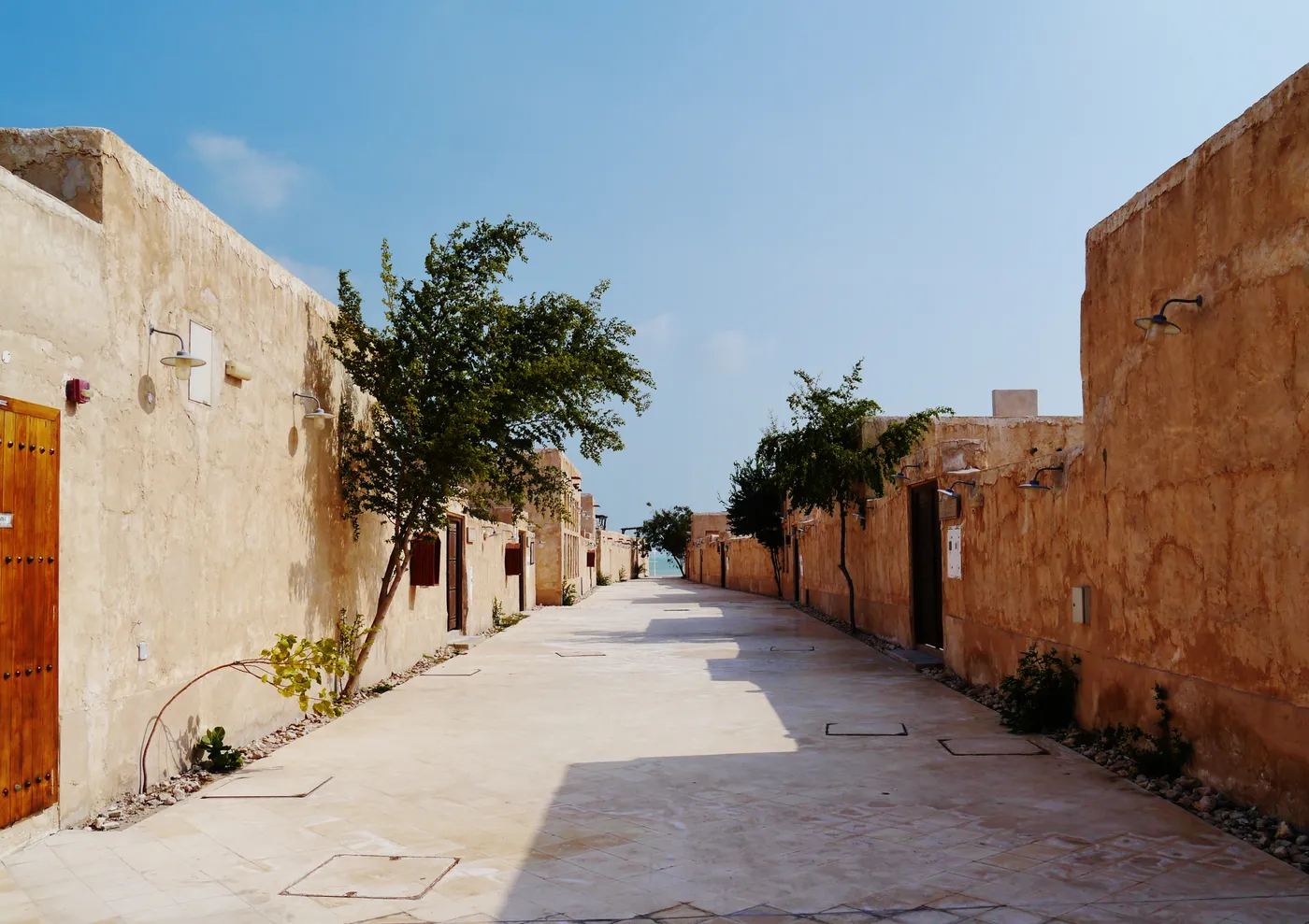 An empty sandstone lane in the old souq at Al Wakrah, plain rendered walls and a heavy timber door on one side, a single tree leaning over the paving and the sea visible at the far end