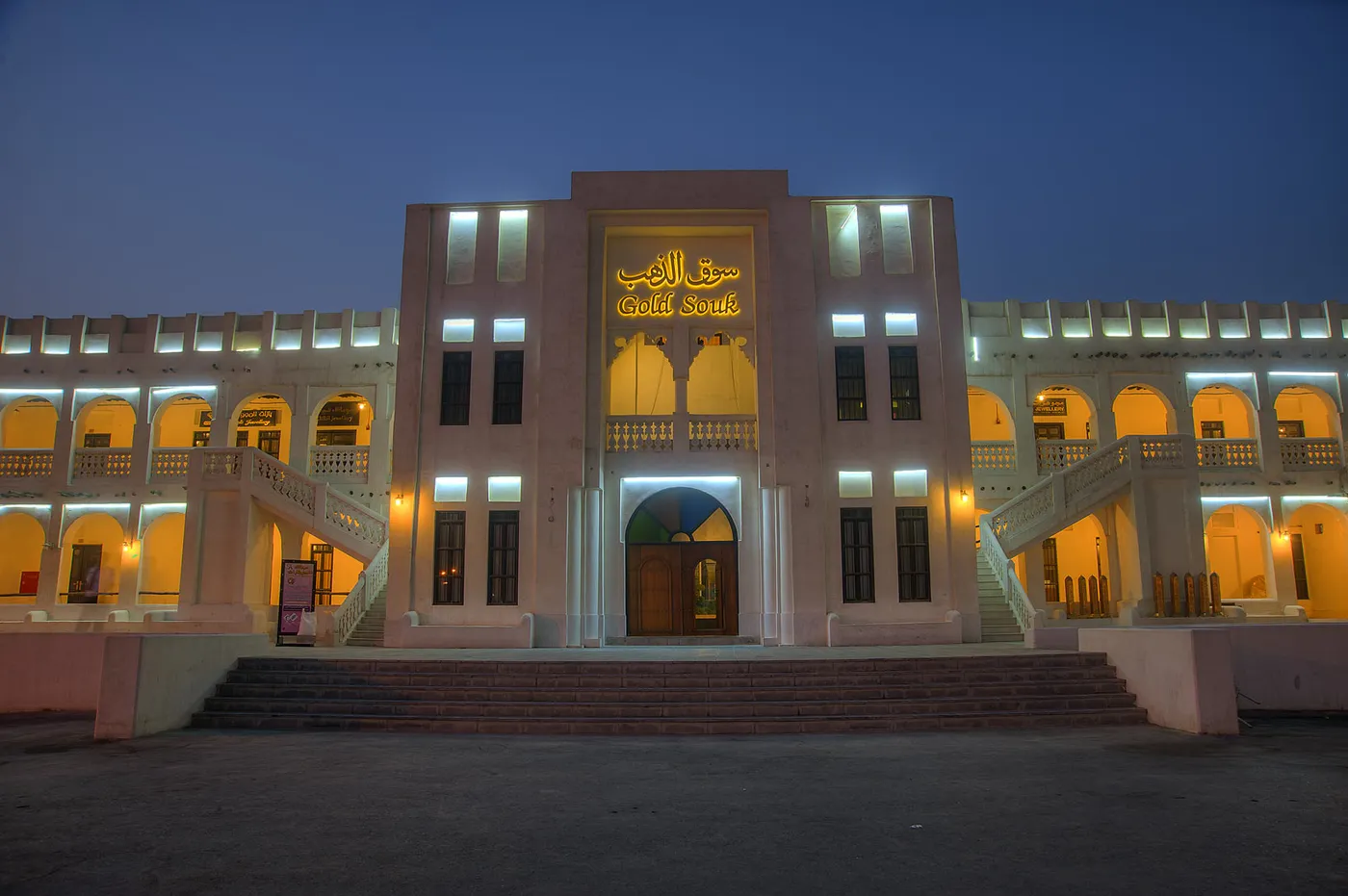 The Gold Souq building at Souq Waqif in Doha lit up at night, a symmetrical two-storey arcade with external staircases and an illuminated sign above the central doorway