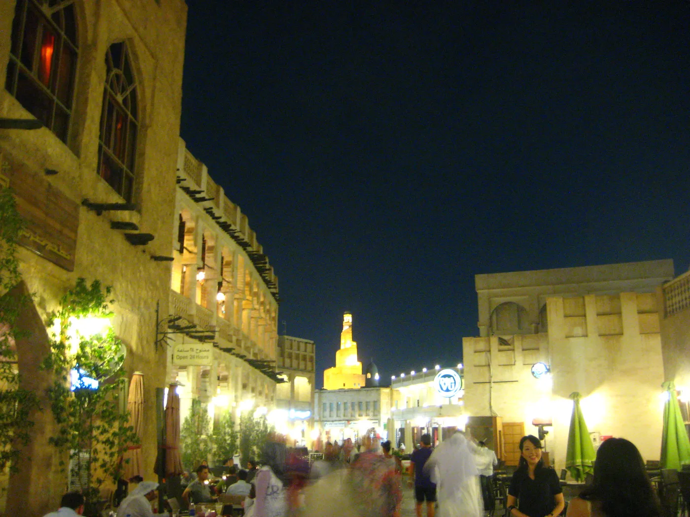The main street of Souq Waqif in Doha after dark, restaurant tables under awnings on both sides, crowds walking the middle and the floodlit spiral minaret at the far end