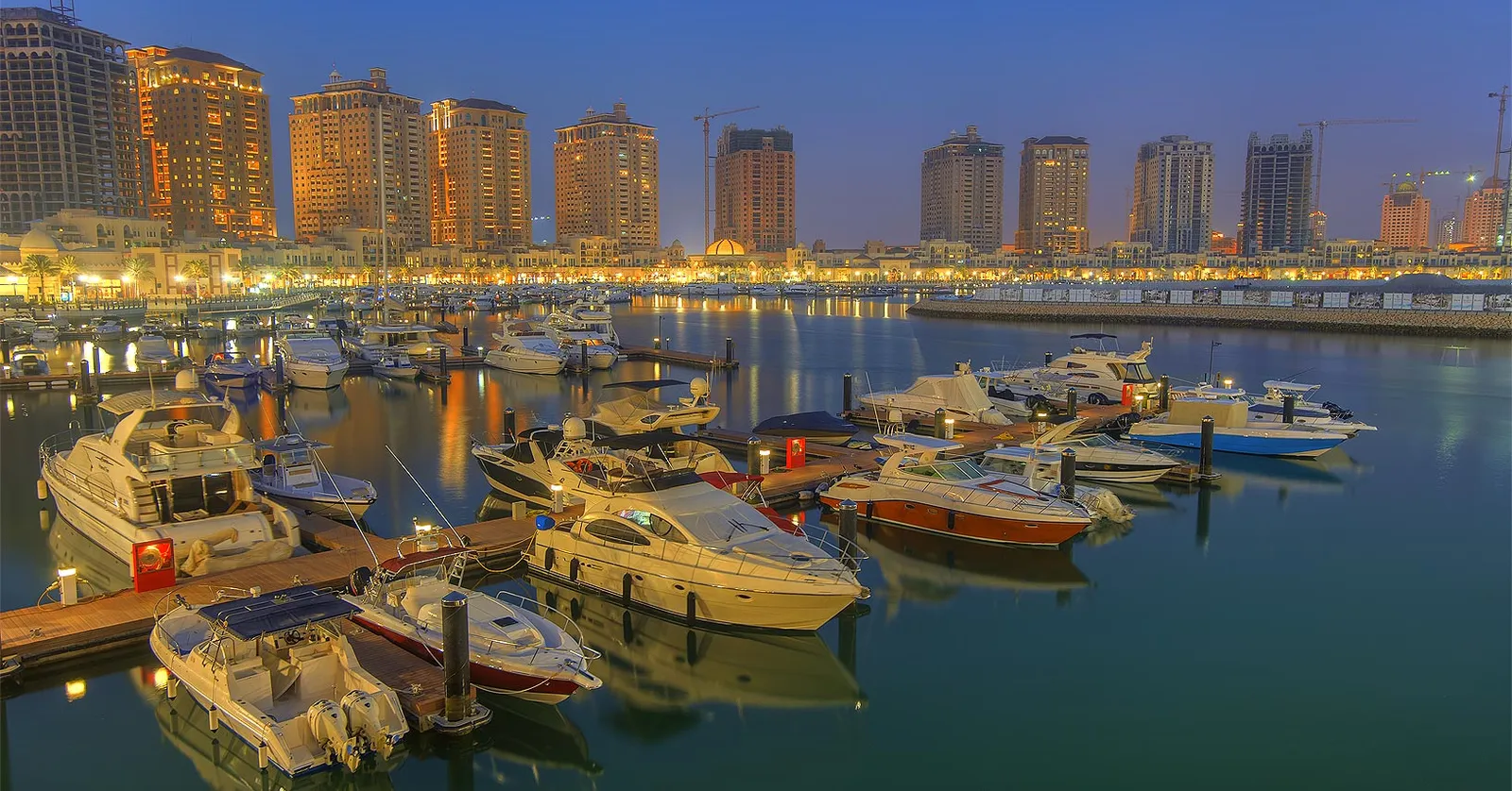 Yachts moored in rows at the Porto Arabia marina on The Pearl-Qatar at dusk, with a curve of lit residential towers behind them reflected in flat water