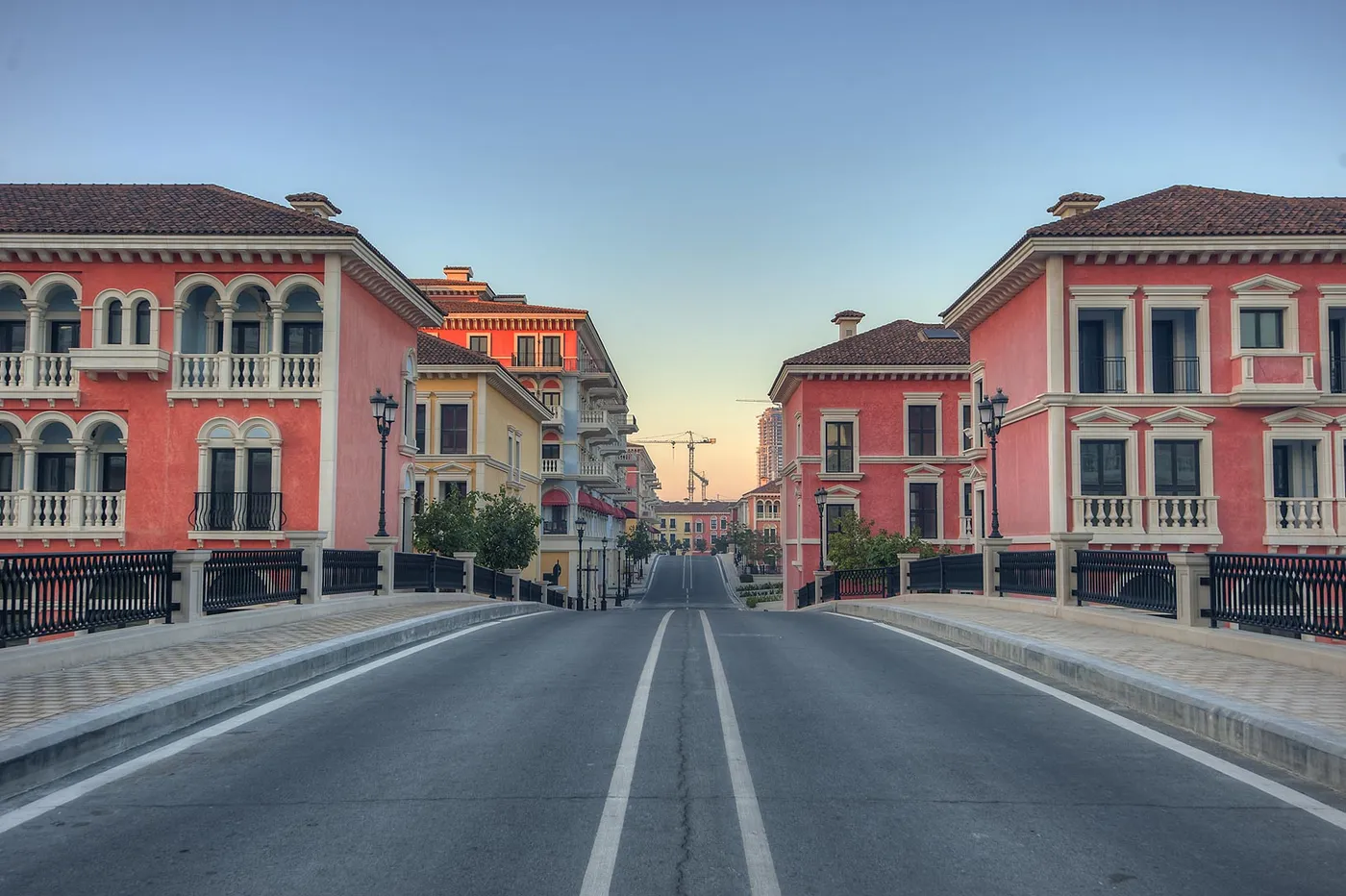 An empty road crossing a bridge between tall pink buildings with balconies and tiled roofs at Qanat Quartier on The Pearl-Qatar, early morning light behind