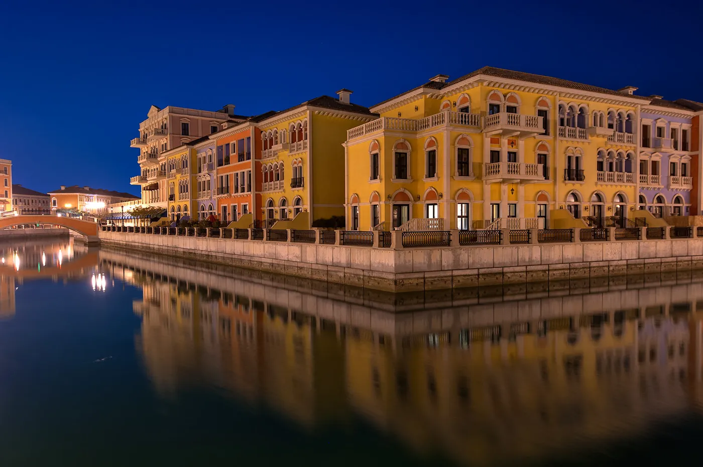 A canal at Qanat Quartier on The Pearl-Qatar after dark, a terrace of yellow, orange and cream Venetian-style buildings along the quay reflected in still water