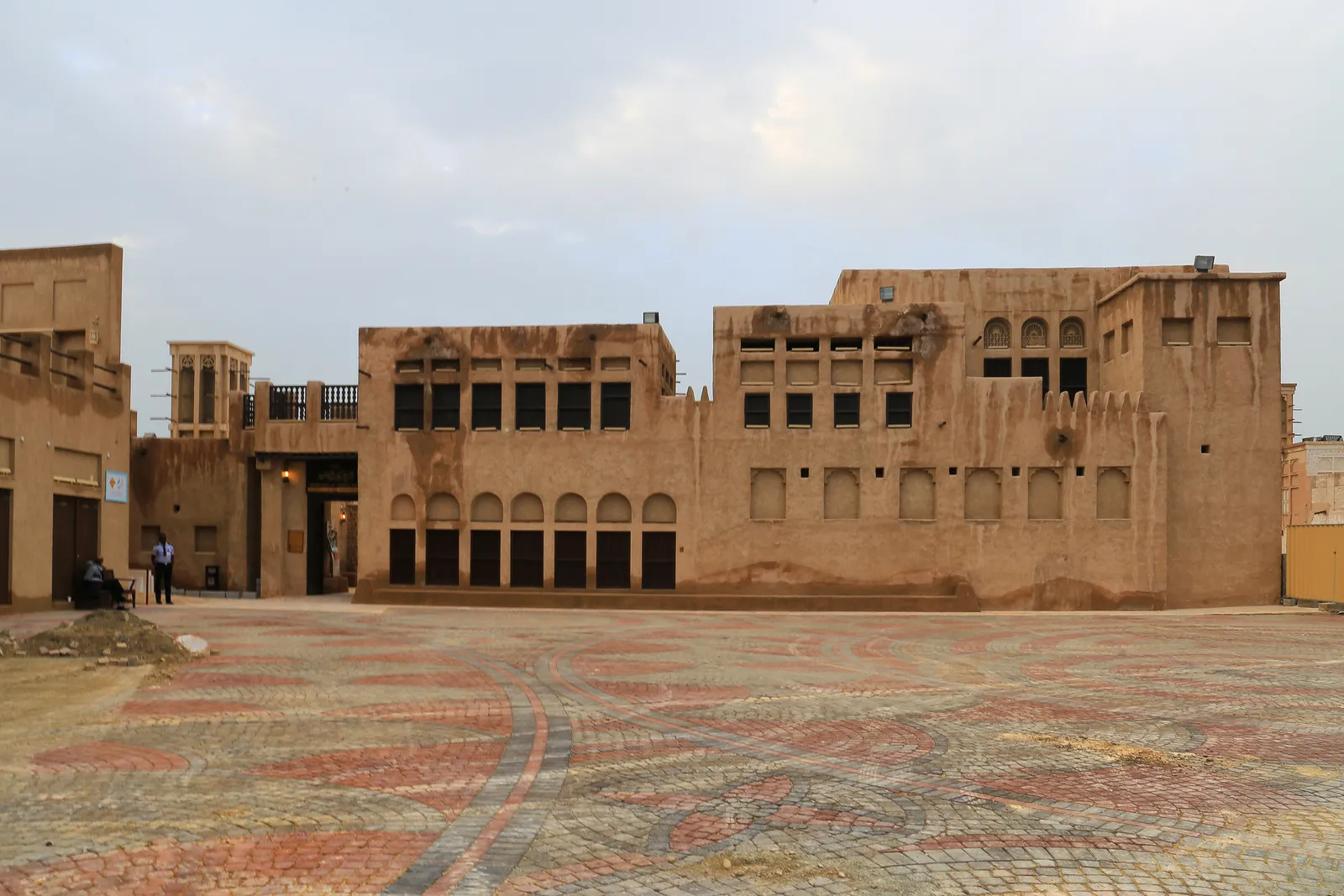 The long sand-coloured facade of Sheikh Saeed Al Maktoum House in Al Shindagha, Dubai, with rows of arched windows and a small wind tower behind a patterned paved square
