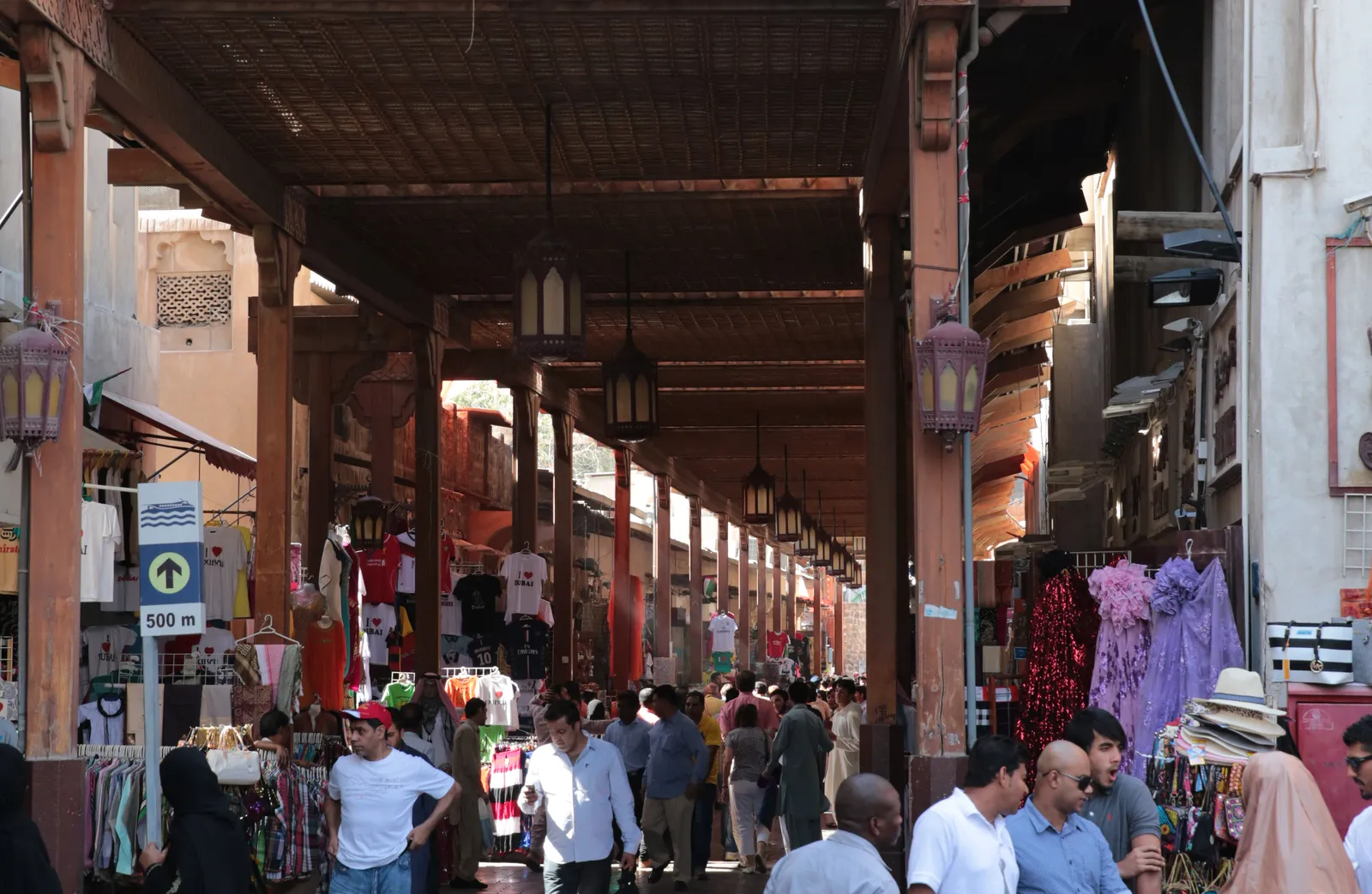 The covered lane of the Textile Souk in Bur Dubai, a wooden roof on carved pillars and hanging lanterns over shops of scarves and clothing, busy with shoppers