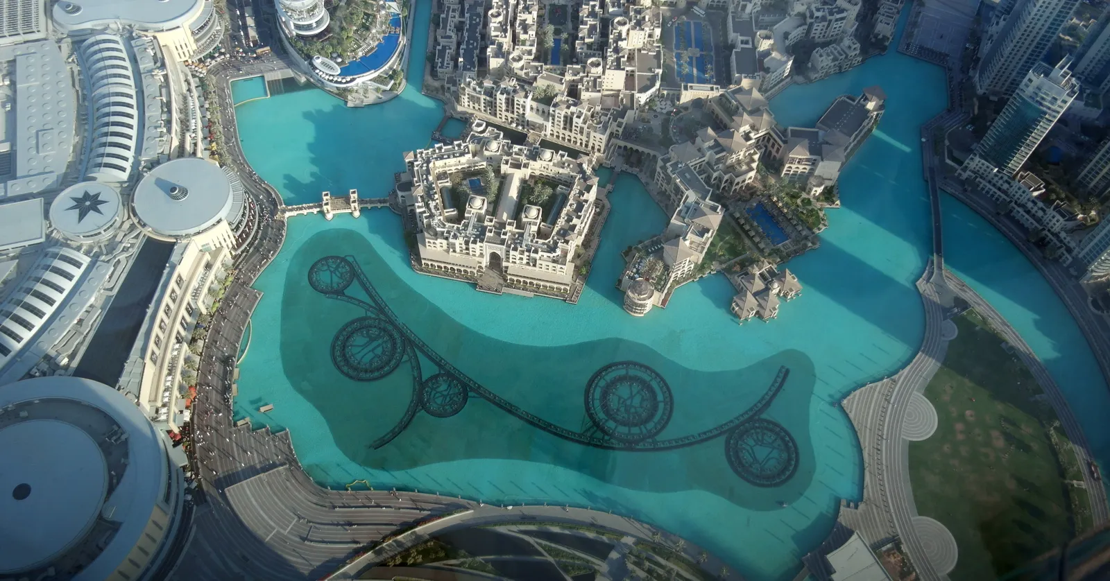 Burj Lake in Downtown Dubai seen from high on Burj Khalifa, turquoise water curving between the domed roofs of Dubai Mall and the low arcaded blocks of Souk Al Bahar