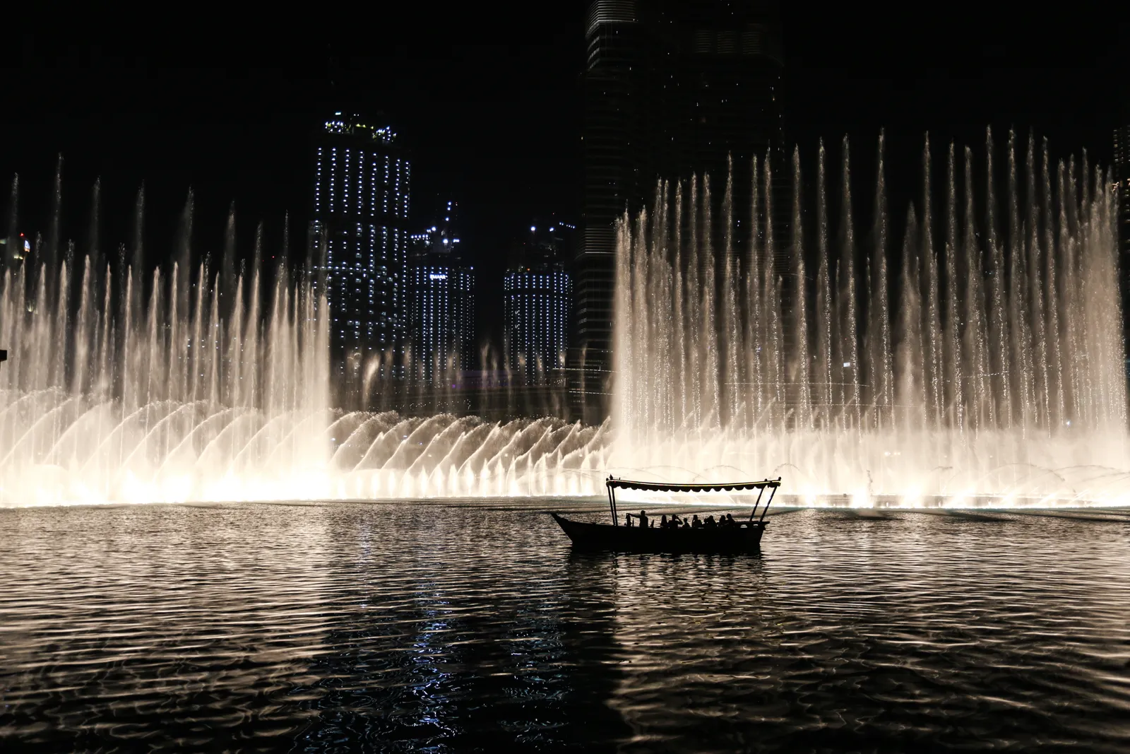 A small wooden abra with a canopy floating on Burj Lake at night in front of a wall of lit fountain jets, with Downtown Dubai's towers behind