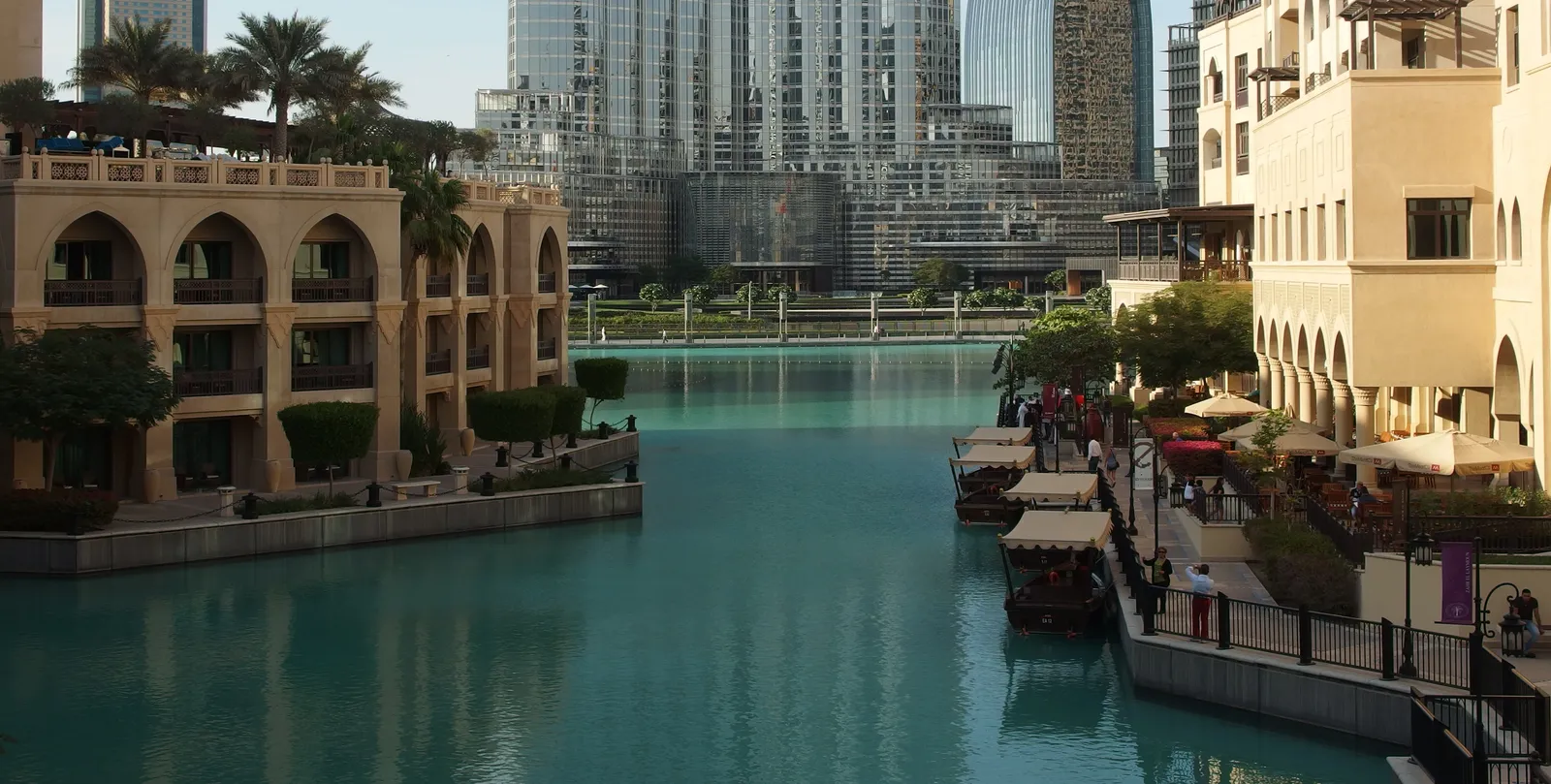 Sand-coloured arcades and balconies of Souk Al Bahar in Downtown Dubai lining a turquoise channel of Burj Lake, with cafe tables along the waterside path and towers beyond
