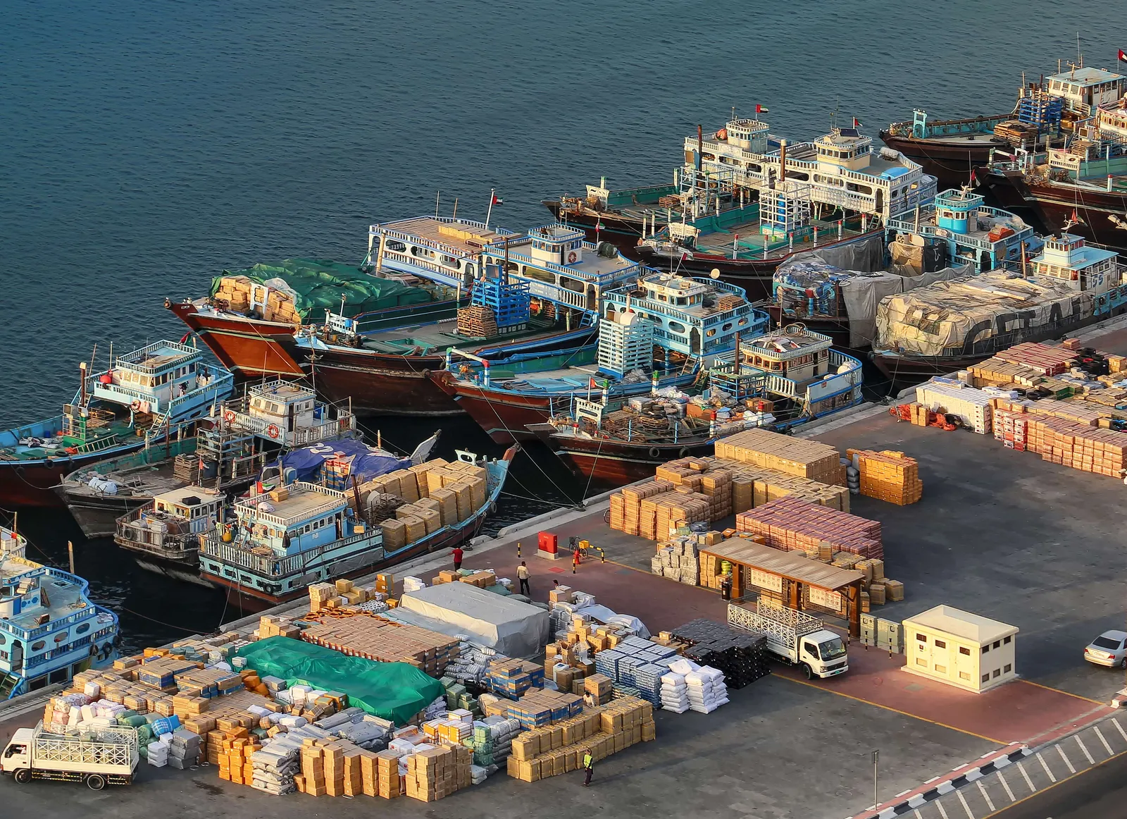 Wooden cargo dhows moored several deep along the Deira wharf on Dubai Creek, seen from above, with pallets and crates of goods stacked on the quay beside them