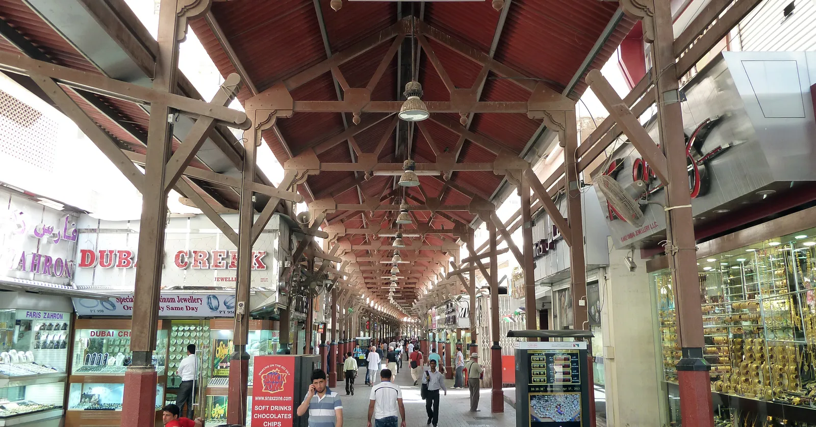 The covered main lane of the Gold Souk in Deira, Dubai, a timber roof on red-painted posts over a paved walkway lined with jewellers' windows full of gold