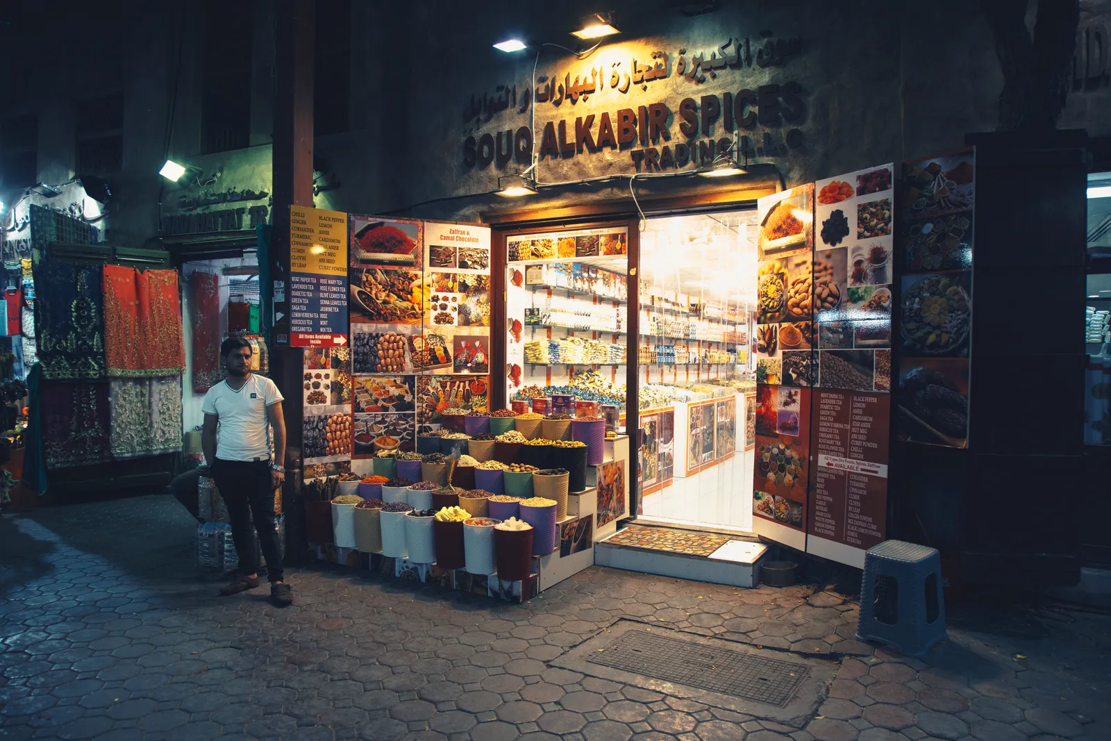 A spice shop in Deira's Spice Souk in Dubai at night, open tubs of dried spices, flowers and herbs stacked outside a lit doorway under an Arabic and English sign