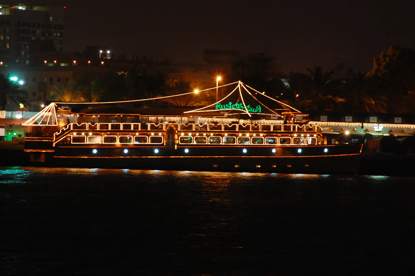 A wooden dhow converted into a floating restaurant, its decks and rigging outlined in strings of lights, moored on Dubai Creek at night