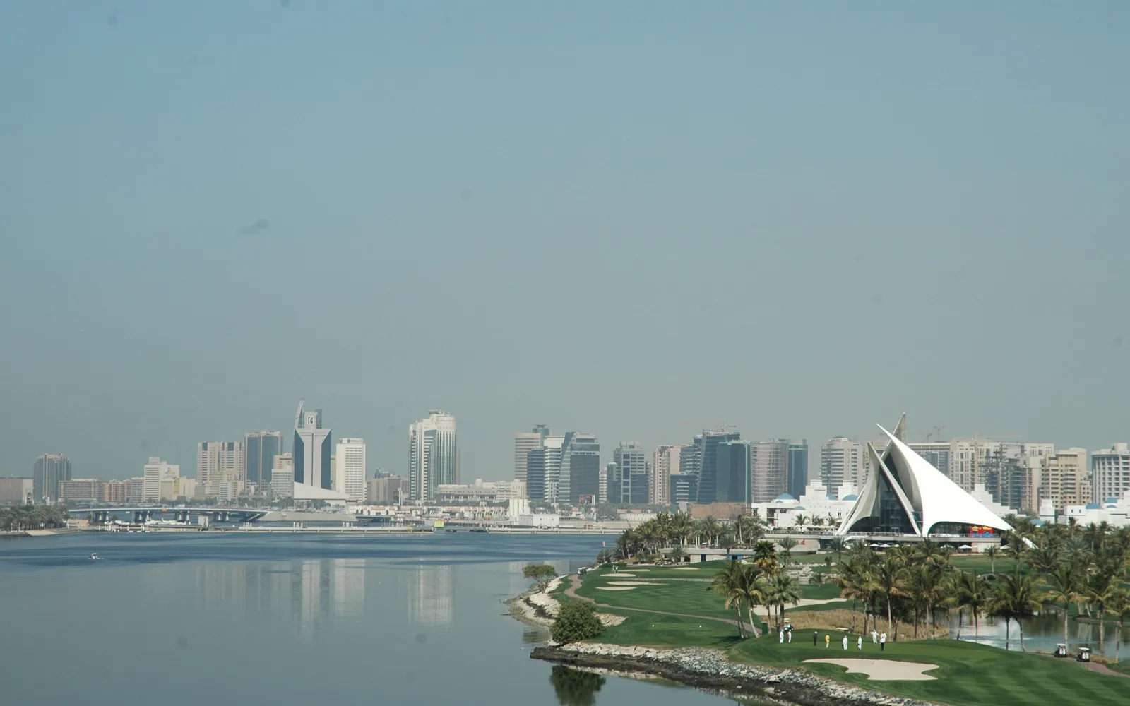 The white peaked clubhouse of Dubai Creek Golf and Yacht Club, shaped like a set of dhow sails, standing among palms and fairways on the edge of Dubai Creek with Deira's towers behind