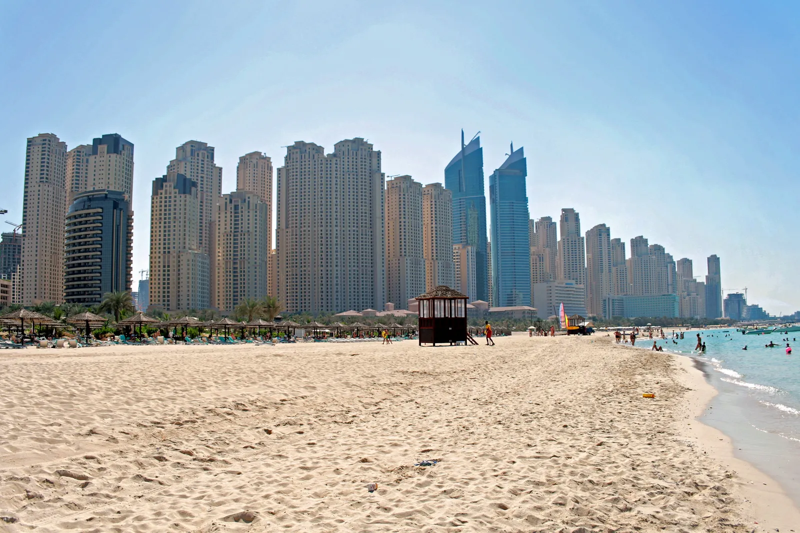 The wide sand of the public beach at Jumeirah Beach Residence in Dubai, with a lifeguard hut, sunshades and a line of tall residential towers behind the shore