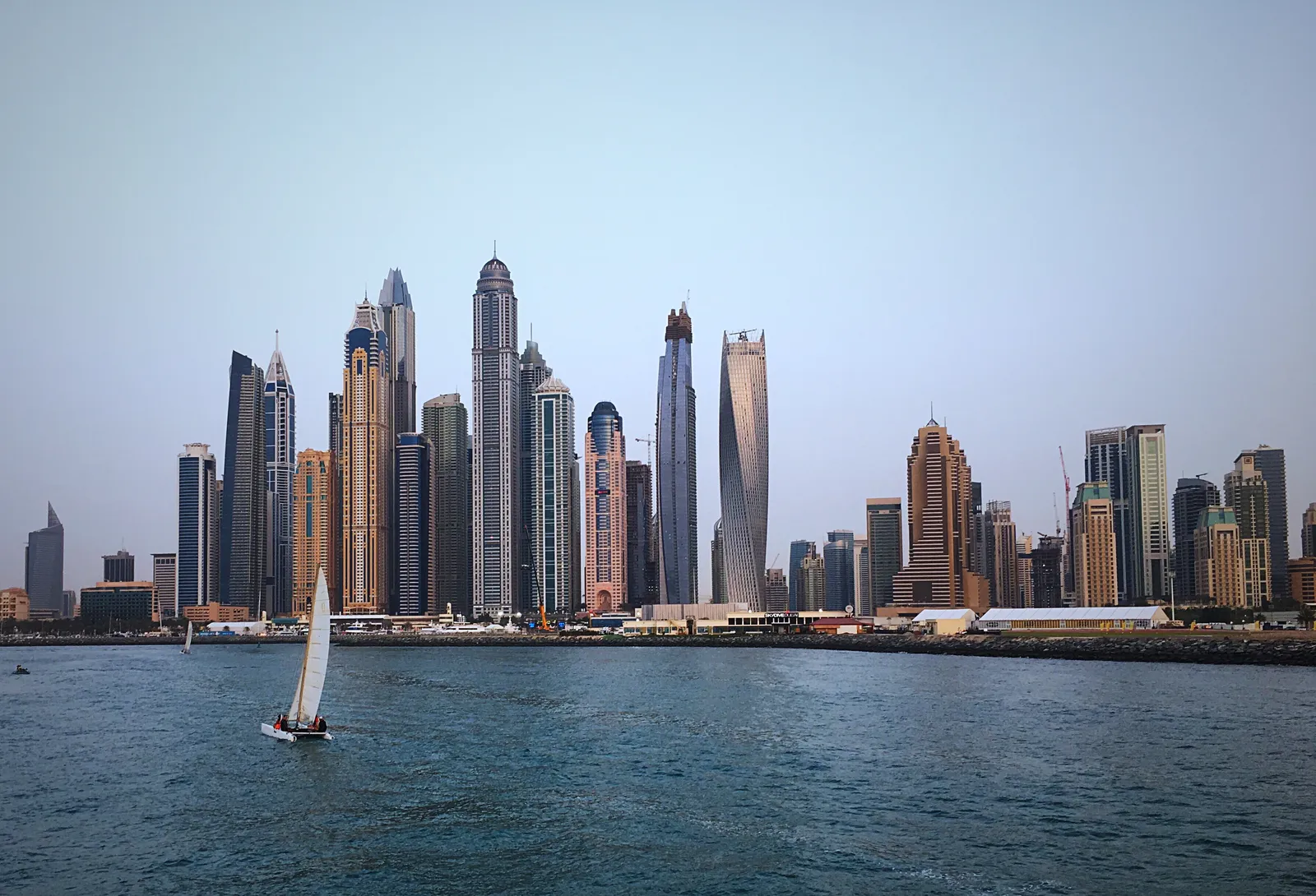 A small sailing boat on the open water off Dubai Marina, with the marina's skyscrapers behind it, including the spiralling Cayan Tower
