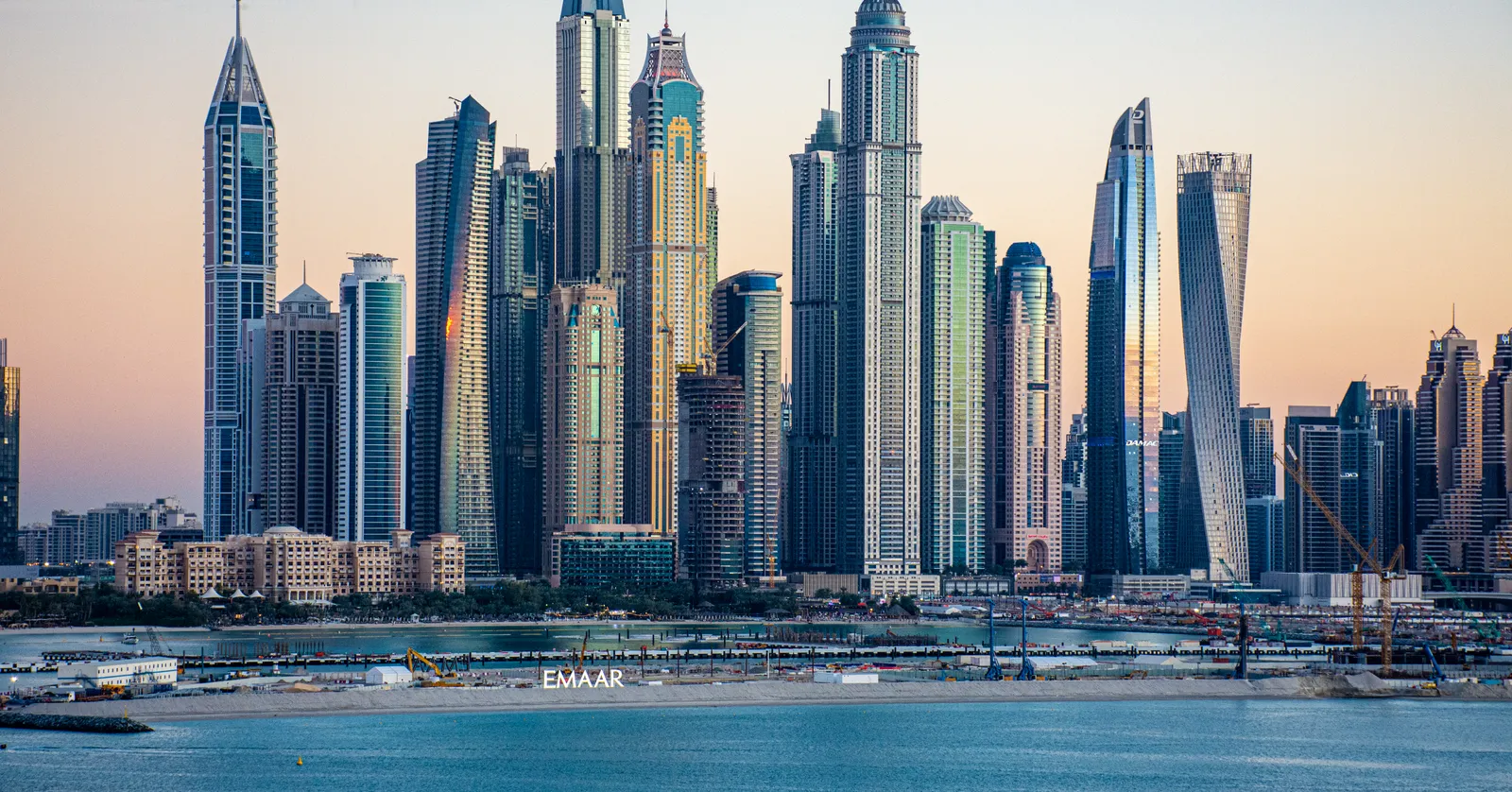 The towers of Dubai Marina at sunset seen from across the water, a tight cluster of skyscrapers with the twisting Cayan Tower on the right