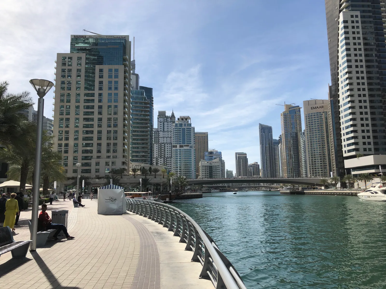 The Marina Walk promenade in Dubai Marina, a curved paved path with benches and a railing along the canal, residential towers on both banks and a footbridge in the distance