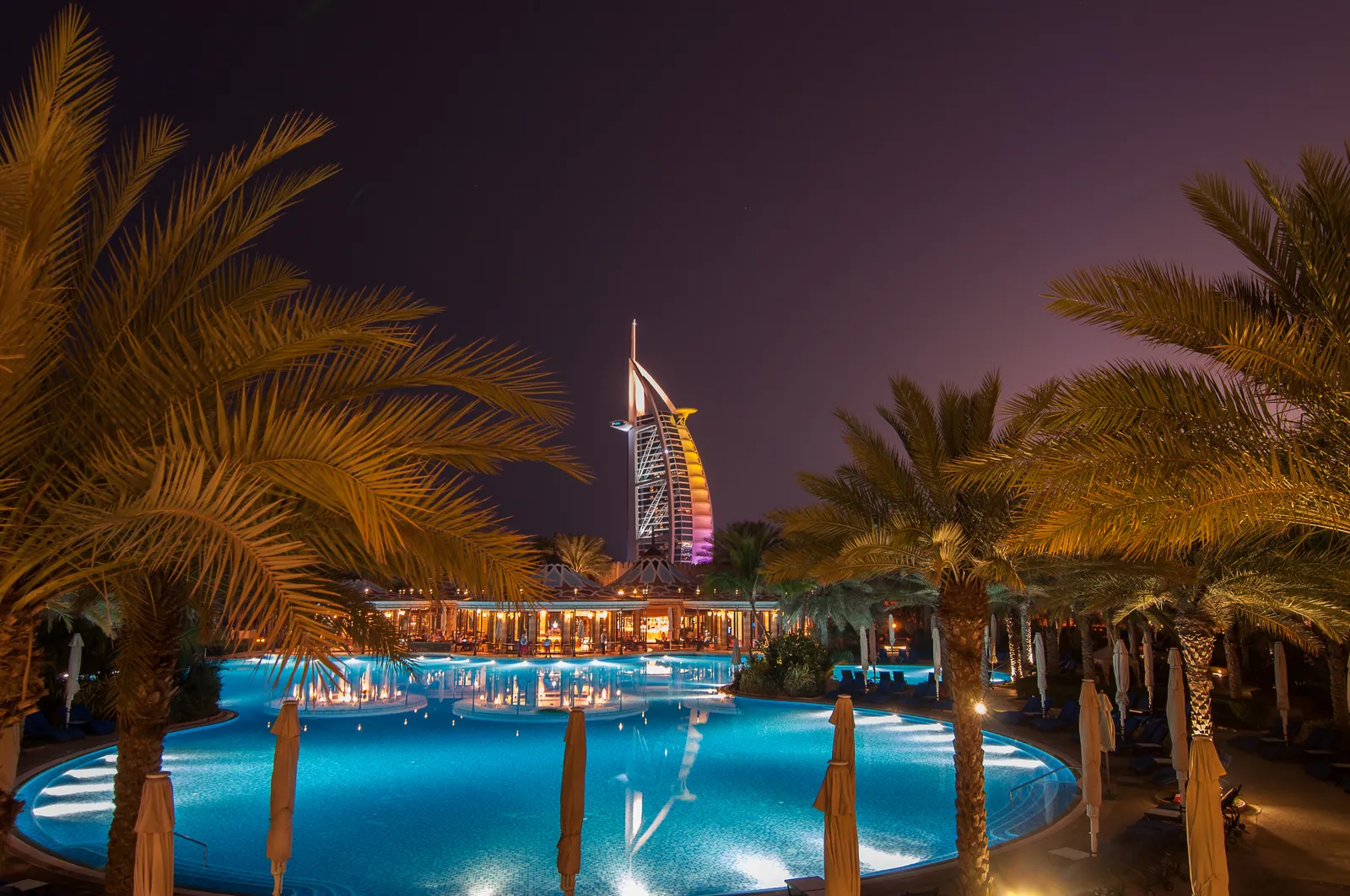 Burj Al Arab lit in shifting colours at night, its sail-shaped tower rising beyond palm trees and a floodlit swimming pool on the Jumeirah shore