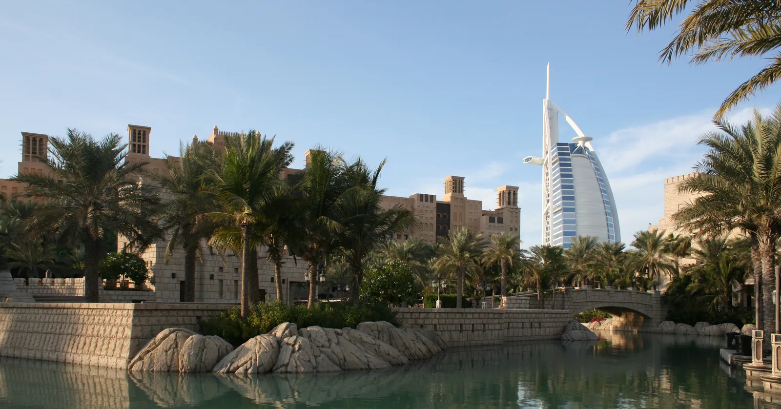 A palm-lined waterway at Madinat Jumeirah in Dubai, with sand-coloured wind-tower buildings on the left and the white sail shape of Burj Al Arab rising behind the trees