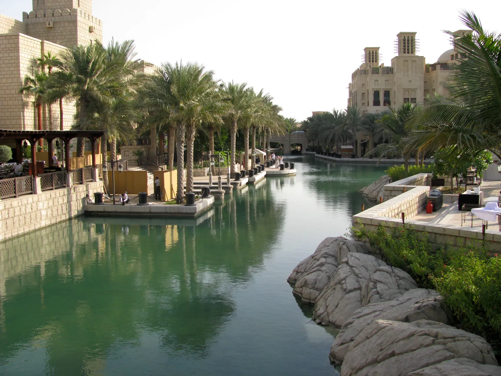 A calm green canal running between palm trees and cafe terraces at Madinat Jumeirah in Dubai, with sand-coloured towers in the style of old Gulf architecture at the far end