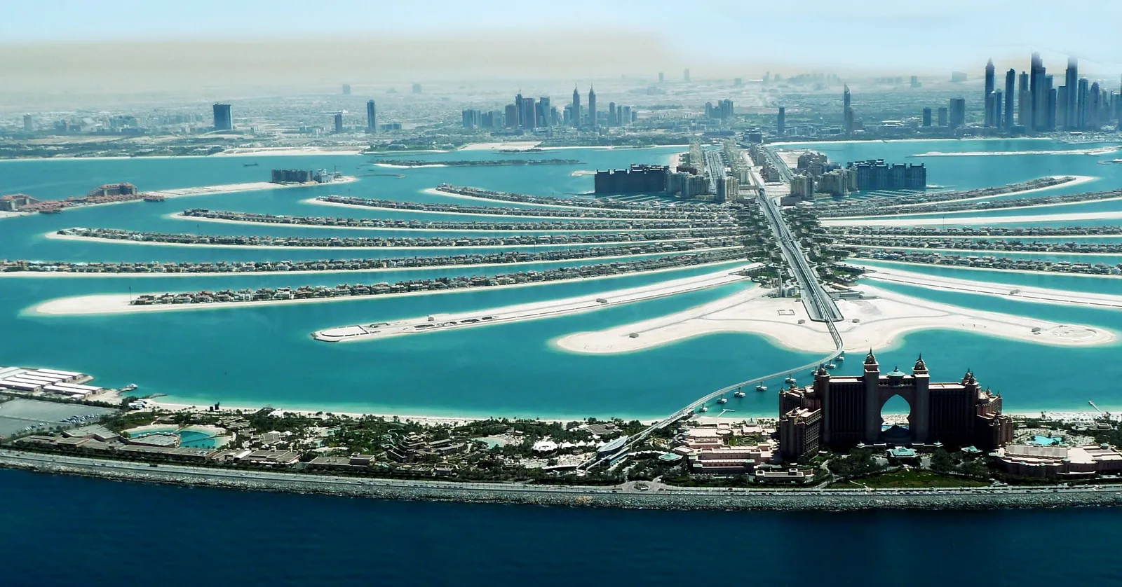 Palm Jumeirah in Dubai from the air, the pink-and-cream Atlantis hotel on the outer crescent in front, the island's sandy fronds and central trunk behind it and the Dubai Marina towers on the far shore